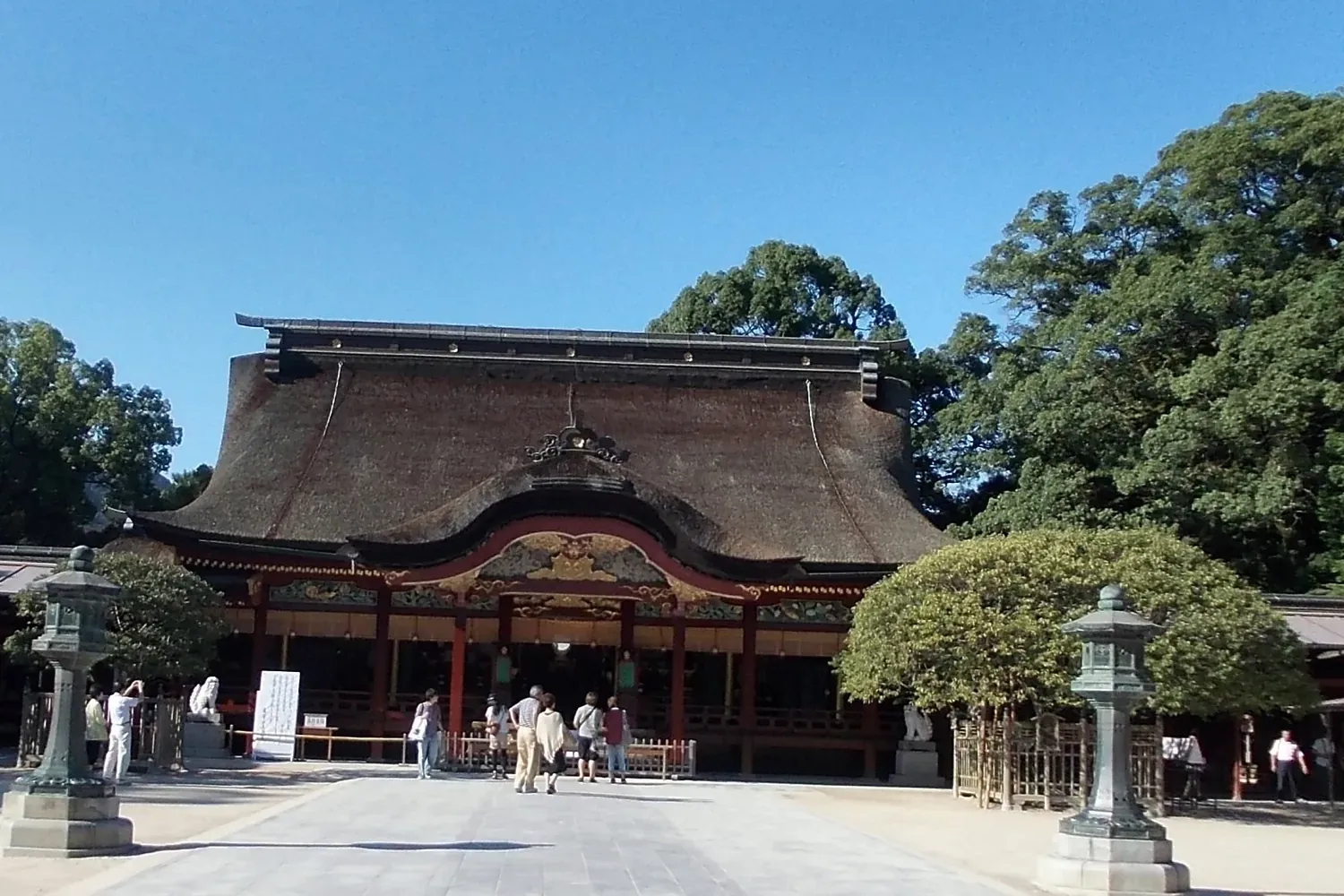 The front of the Dazaifu Tenmangu main hall, a wide red-trimmed building under a deep brown bark roof, with stone lanterns and a few visitors