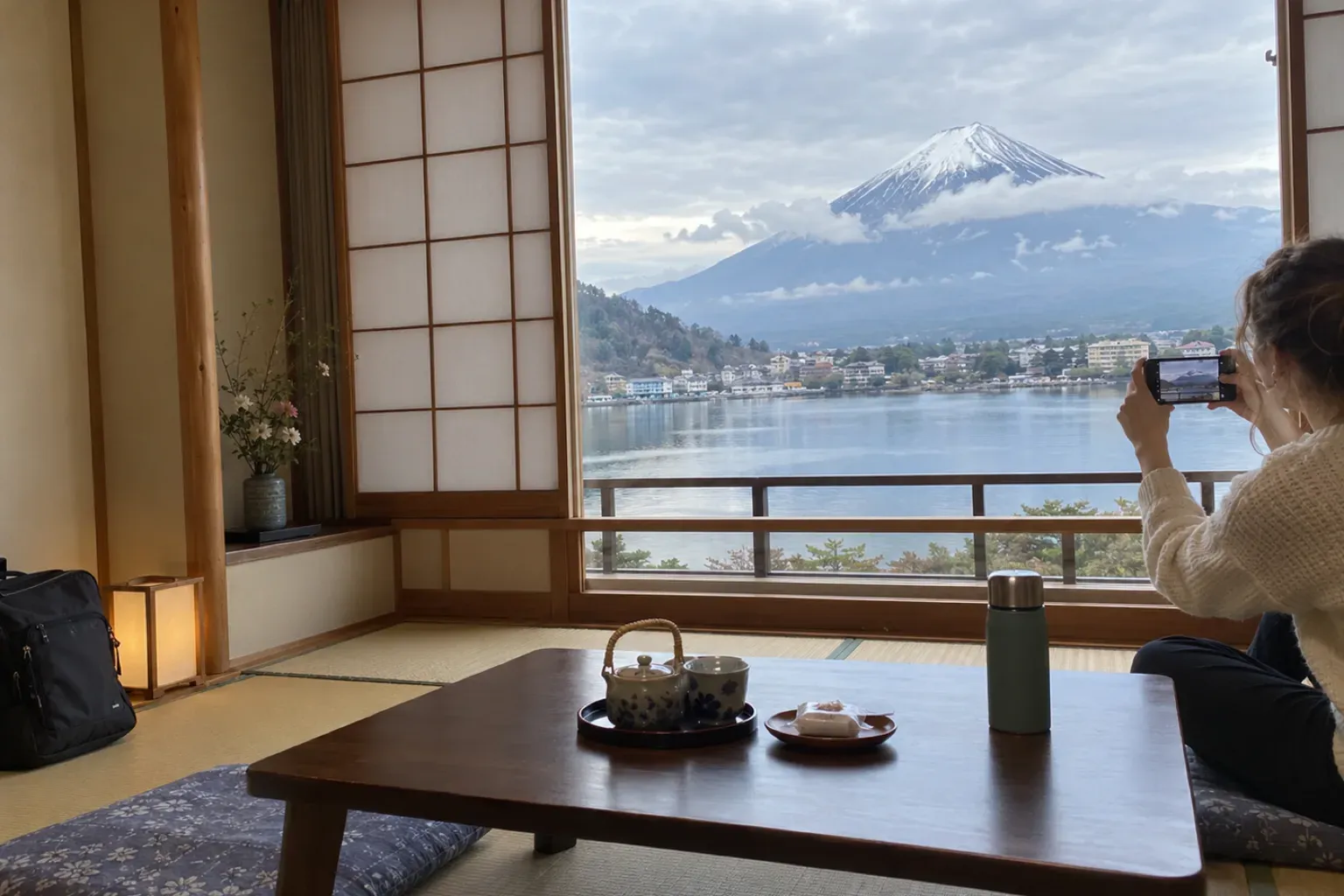A young traveller sits on the tatami by an open window, taking a phone photo of Mount Fuji across Lake Kawaguchi, with a low table and a teapot in front