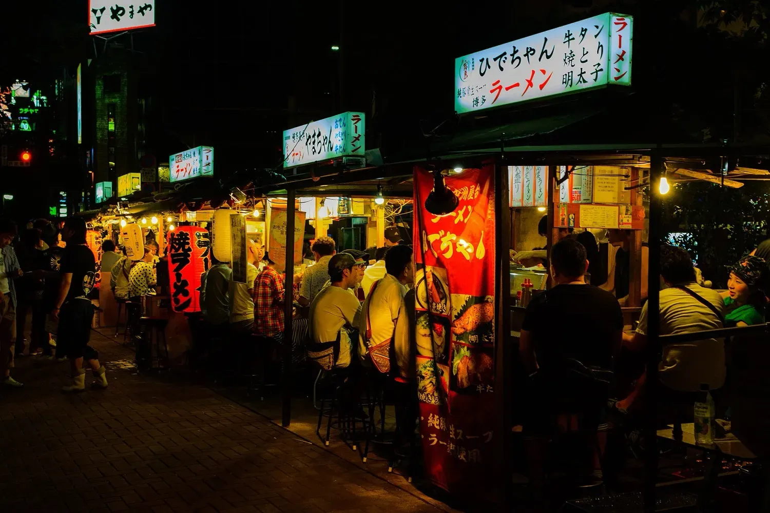 A row of lit yatai stalls in Nakasu at night, with customers on stools and a sign for Hidechan ramen at the right