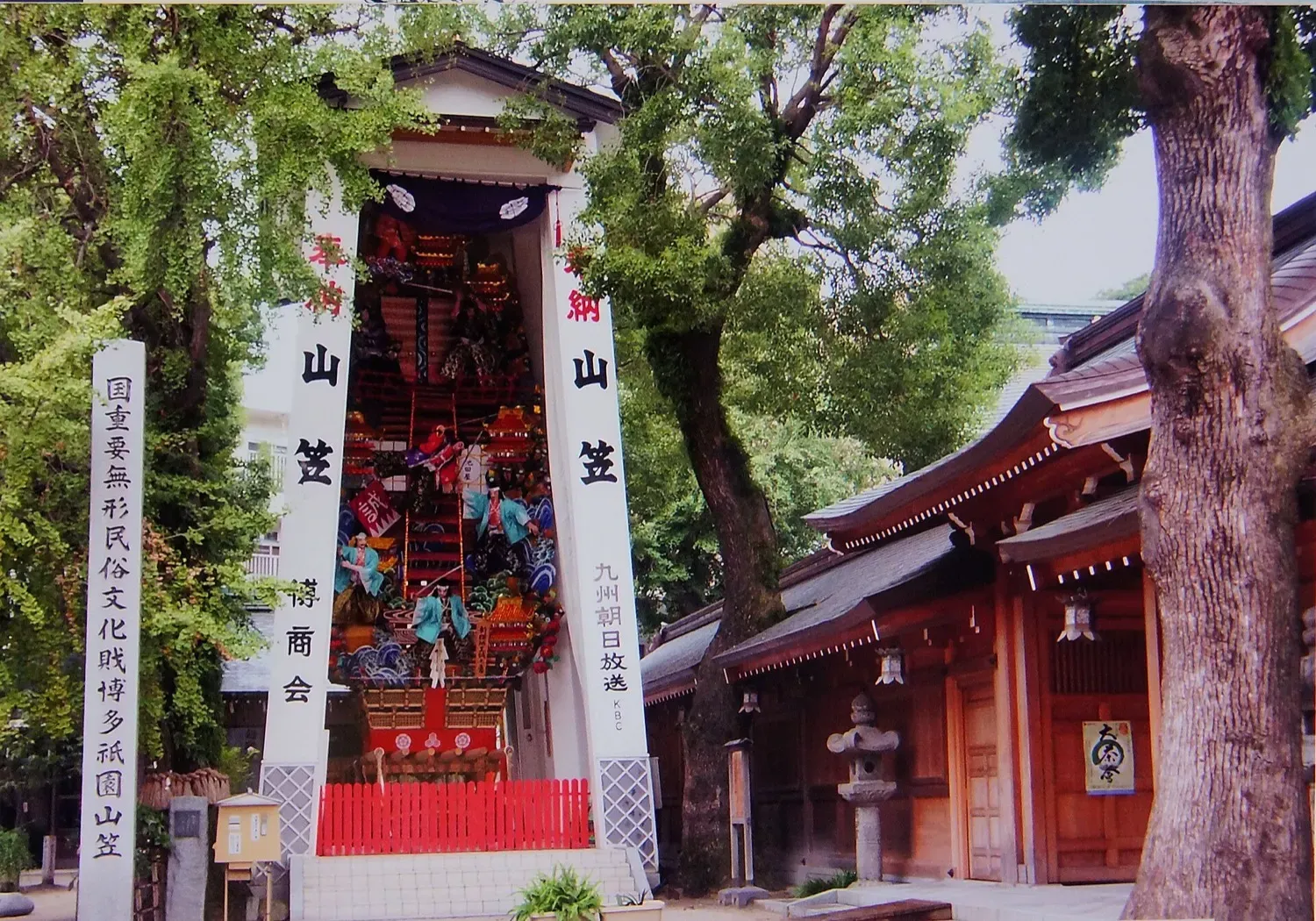 A tall decorated Yamakasa float with warrior figures standing beside a red-trimmed shrine building under trees at Kushida Shrine