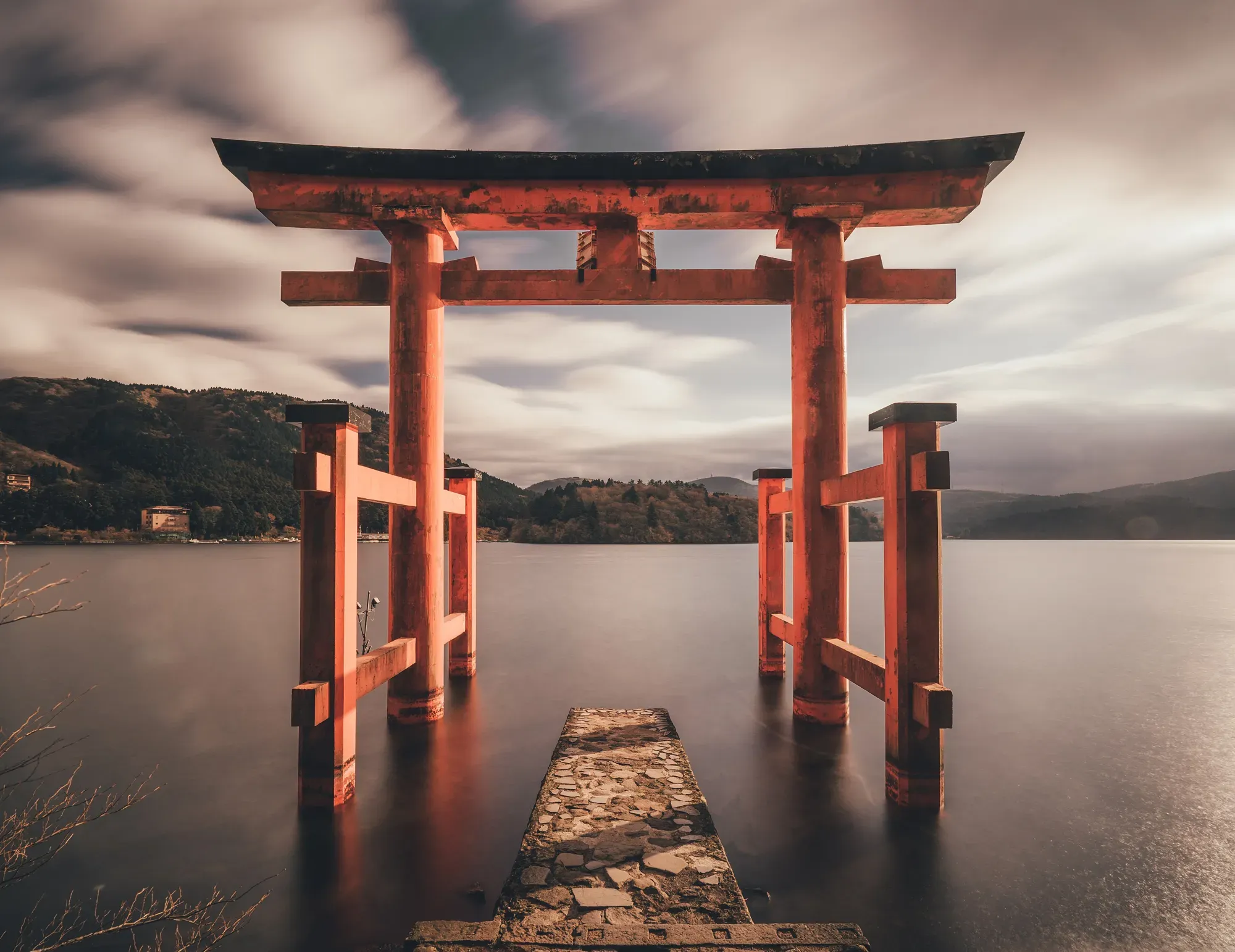 The vermilion gate of Hakone Shrine standing in Lake Ashi, the water smoothed by a long exposure under a cloudy sky