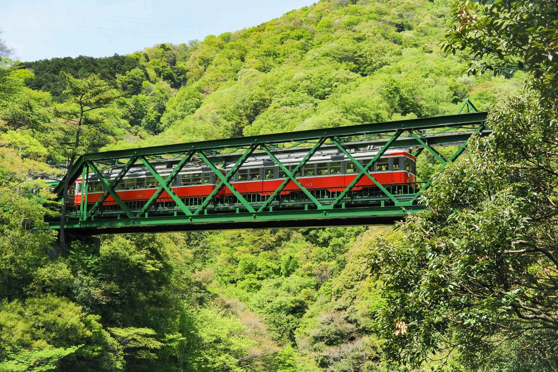 A red-striped Hakone Tozan Railway train crossing the green iron Deyama bridge between wooded hills