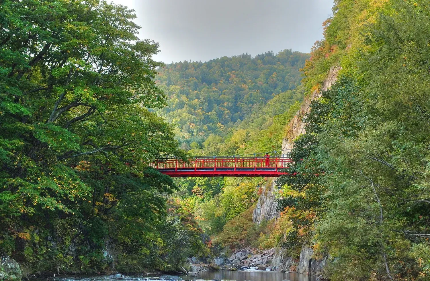 A red suspension bridge over a clear river in a wooded gorge, the leaves just starting to turn