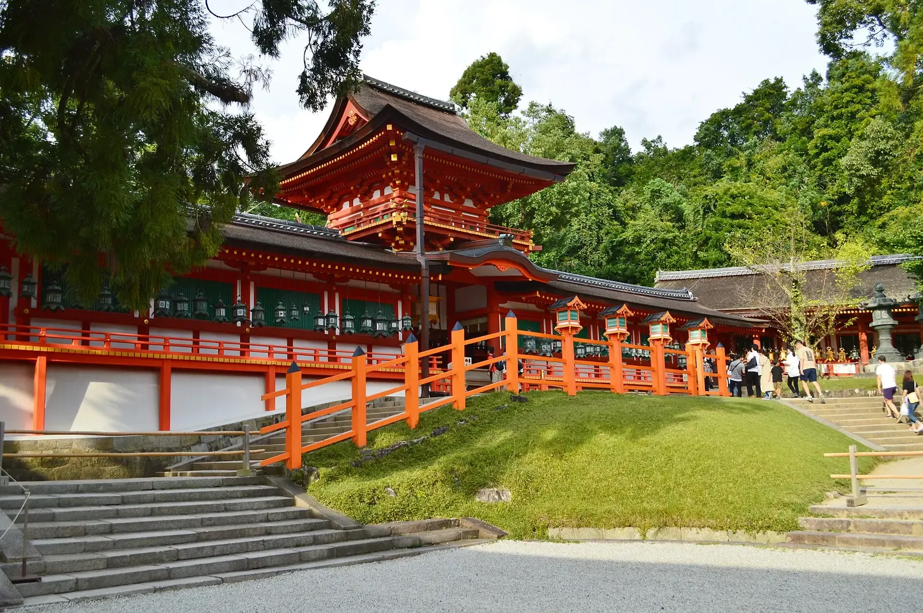 The vermilion middle gate at Kasuga Taisha seen from the approach, with the lantern-hung corridor running to either side and visitors walking past