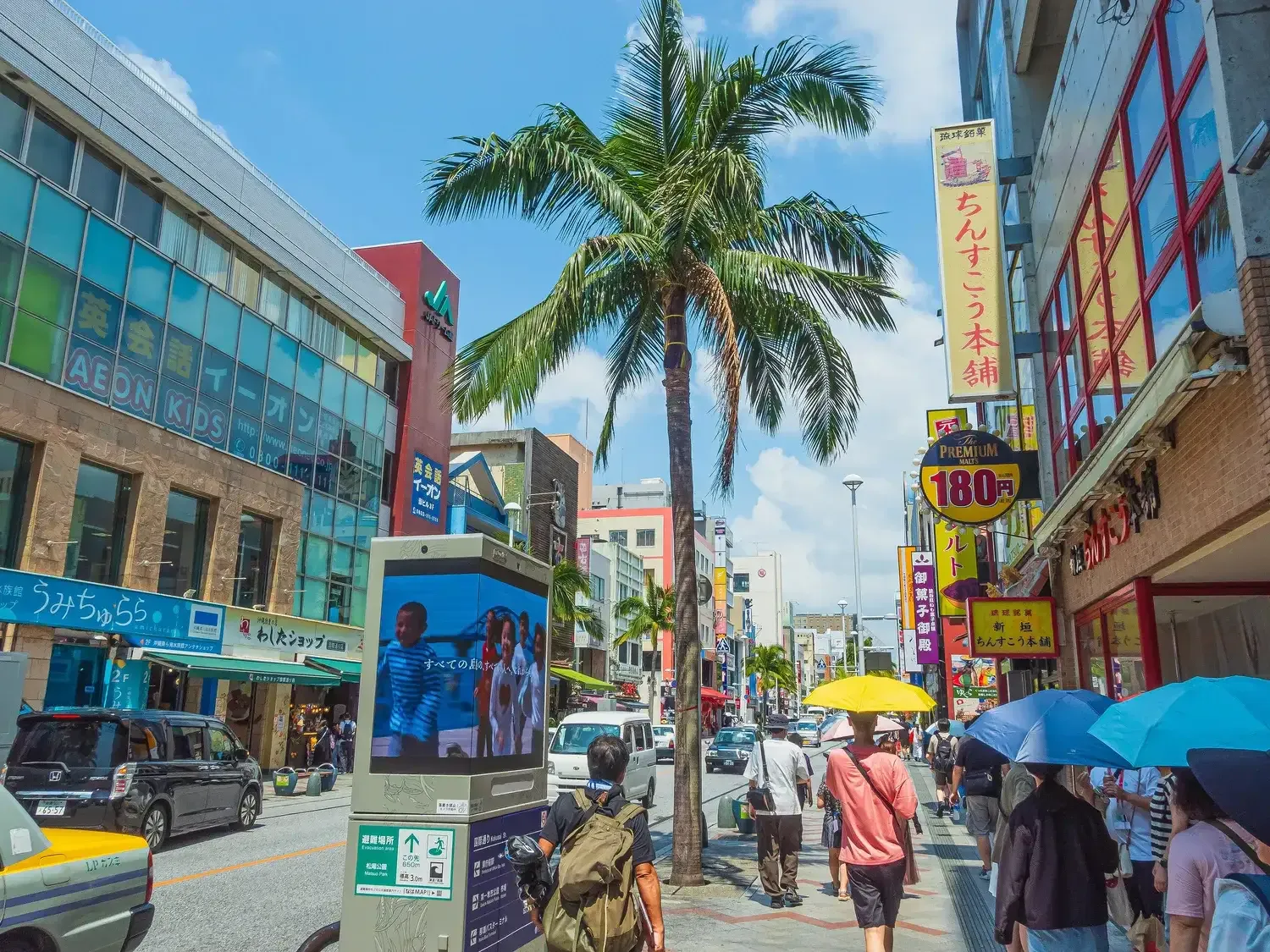 Kokusai Street on a summer day, palm trees and tall shop signs above people walking under umbrellas