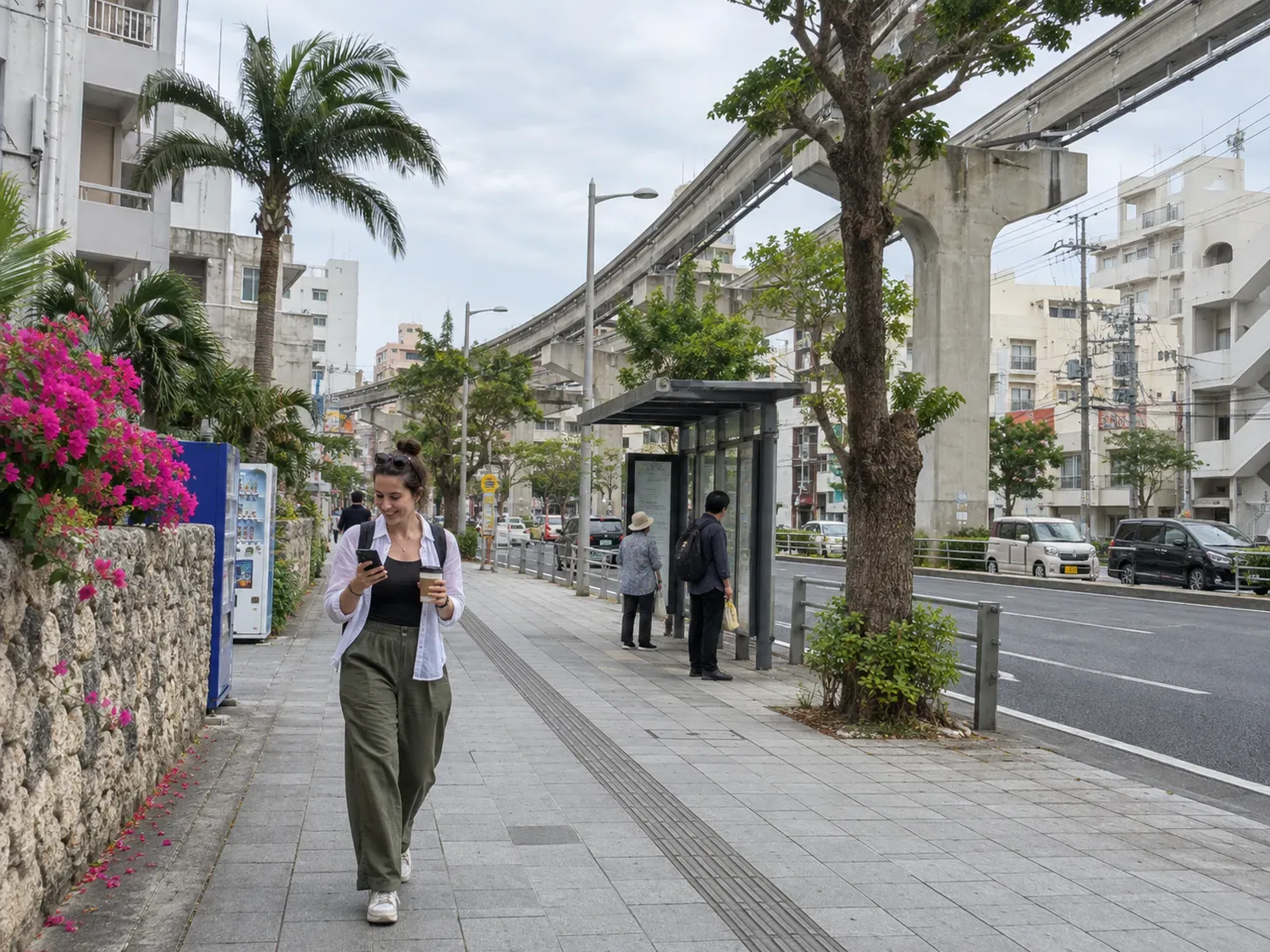 A traveler with a backpack and a takeaway coffee smiling at her phone map as she walks along a sidewalk under the elevated monorail track in Naha on an overcast morning, bougainvillea spilling over a stone wall beside her