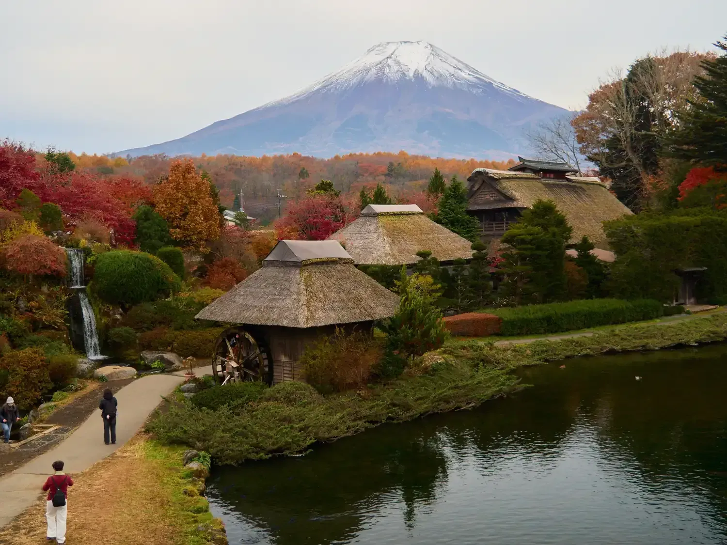 Thatched-roof buildings and a waterwheel beside a pond at Oshino Hakkai, with Mount Fuji behind them in autumn