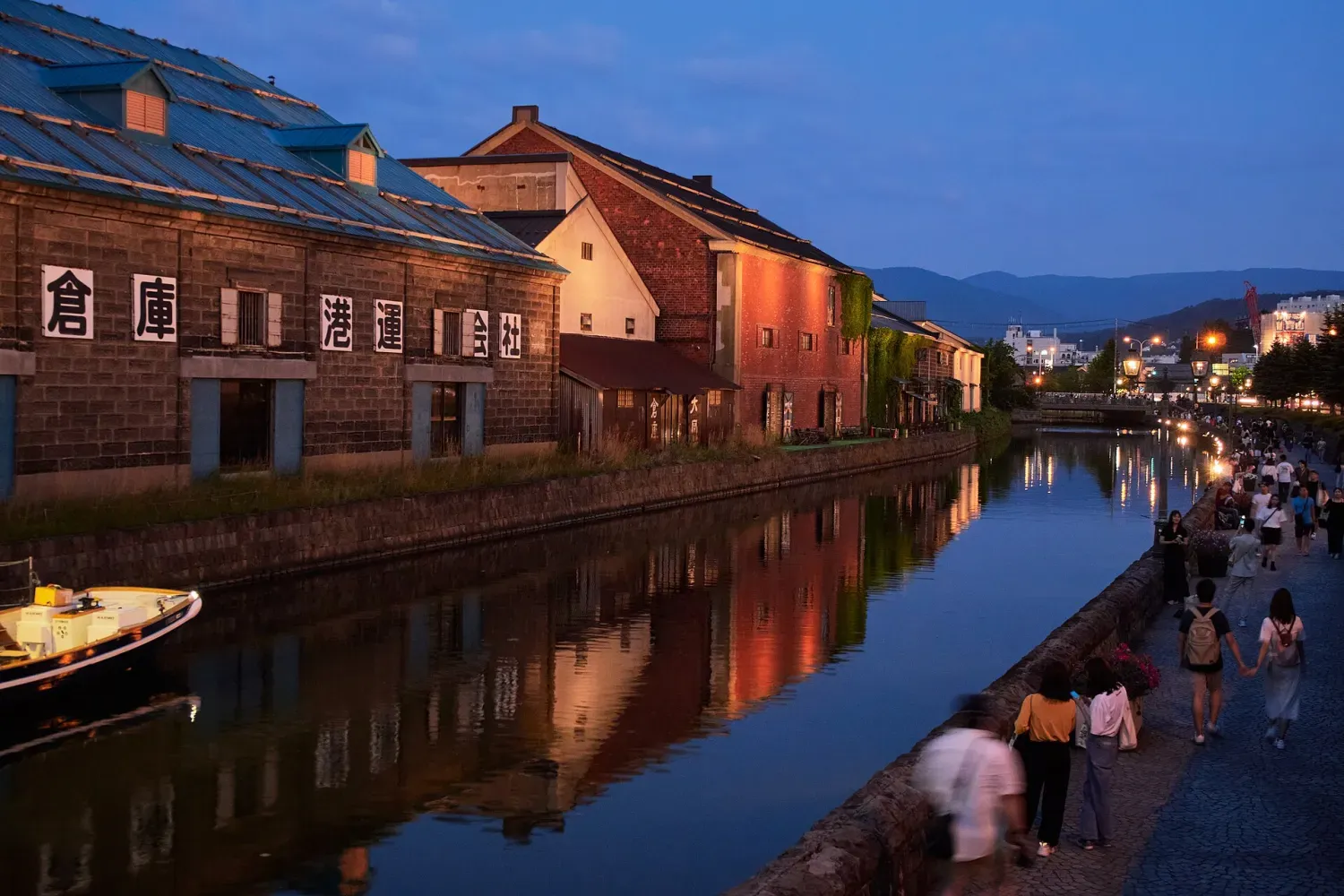 Stone warehouses lit at dusk along a calm canal under a deep blue sky, with people walking on the lamp-lit path