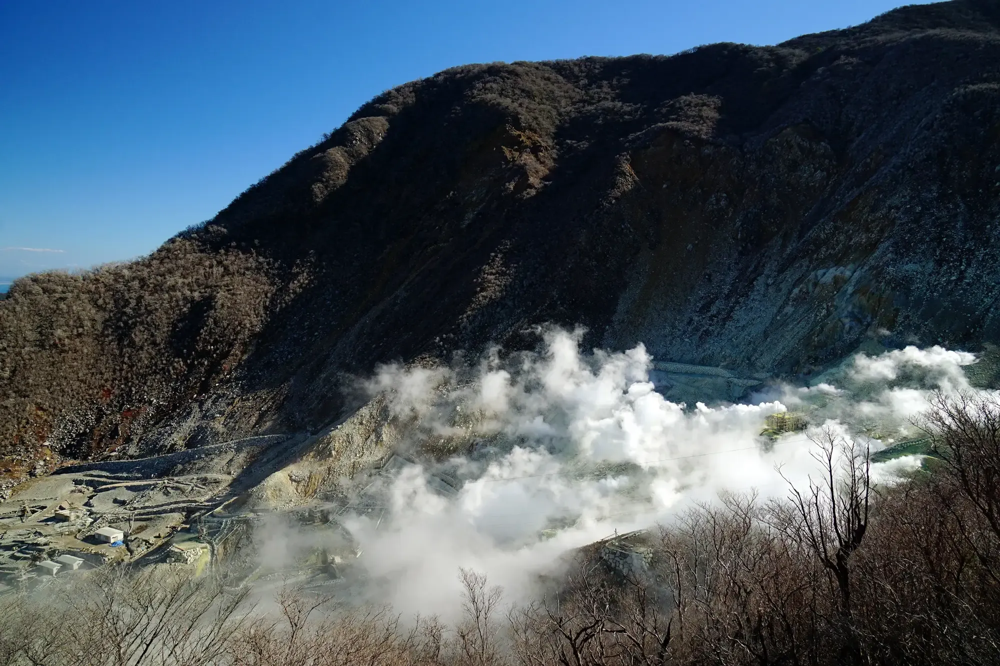 A great plume of white steam rising from the Owakudani valley under a blue sky, below a dark mountainside