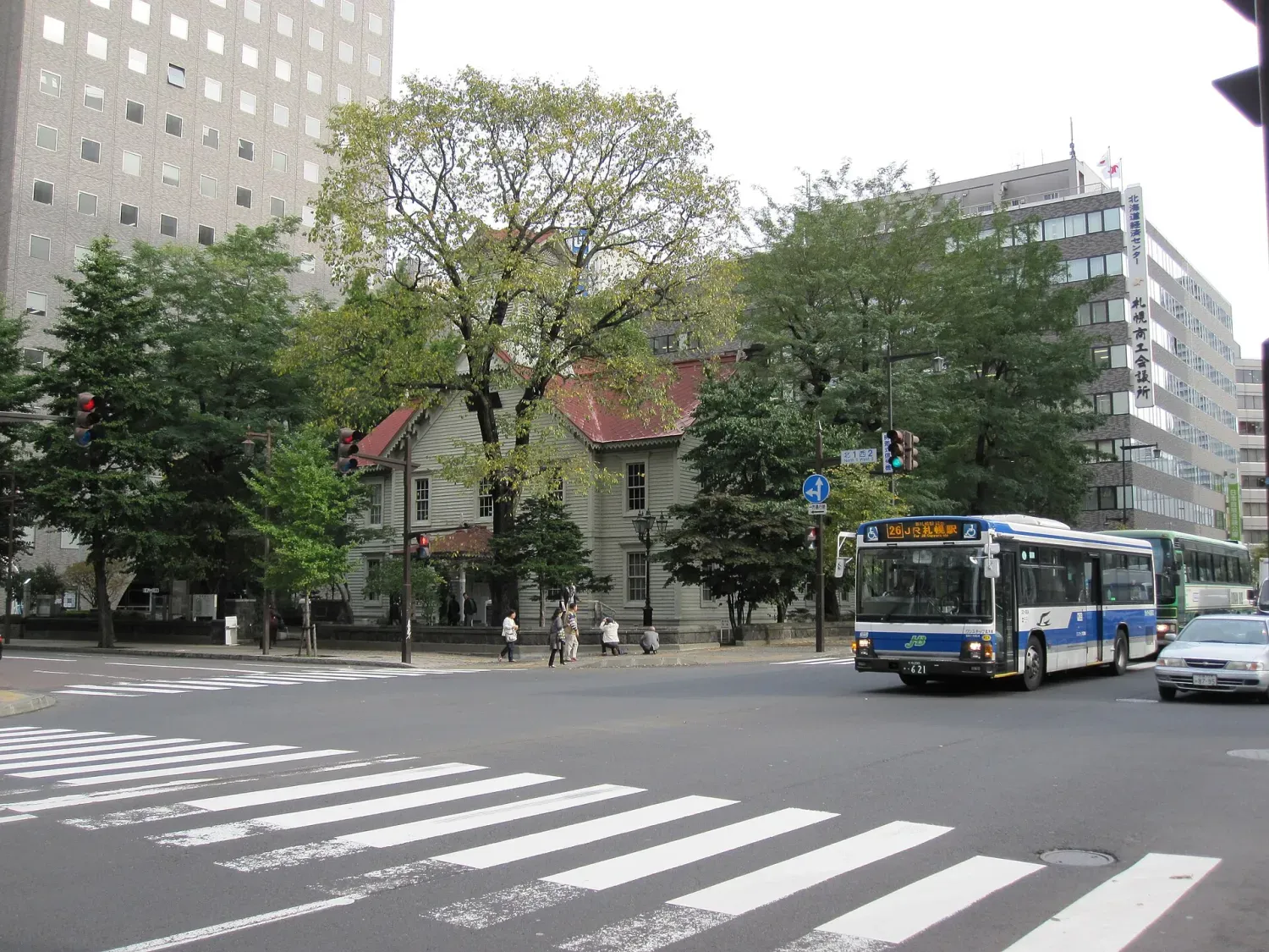 A small wooden hall with a red roof and a clock tower among trees, office blocks rising behind it and a city bus passing