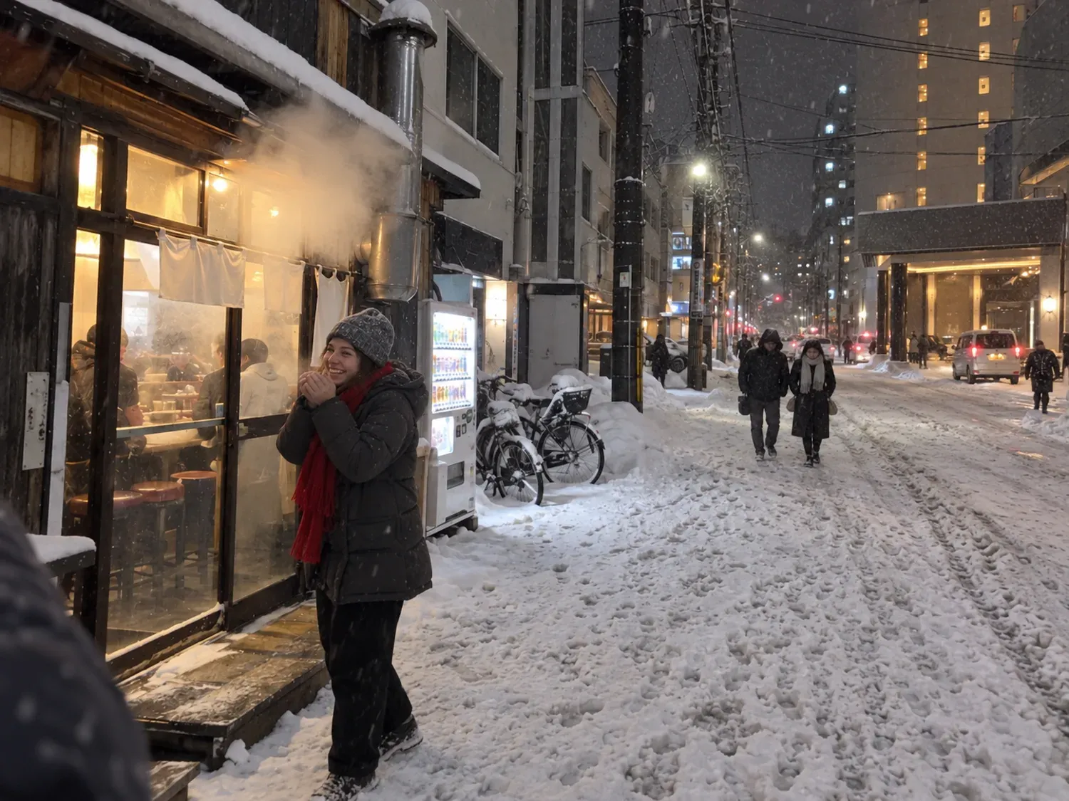 A traveler in a knit hat and red scarf smiling and warming her hands outside a steamy ramen shop on a snowy side street at night, a vending machine and bicycles buried in snow beside her