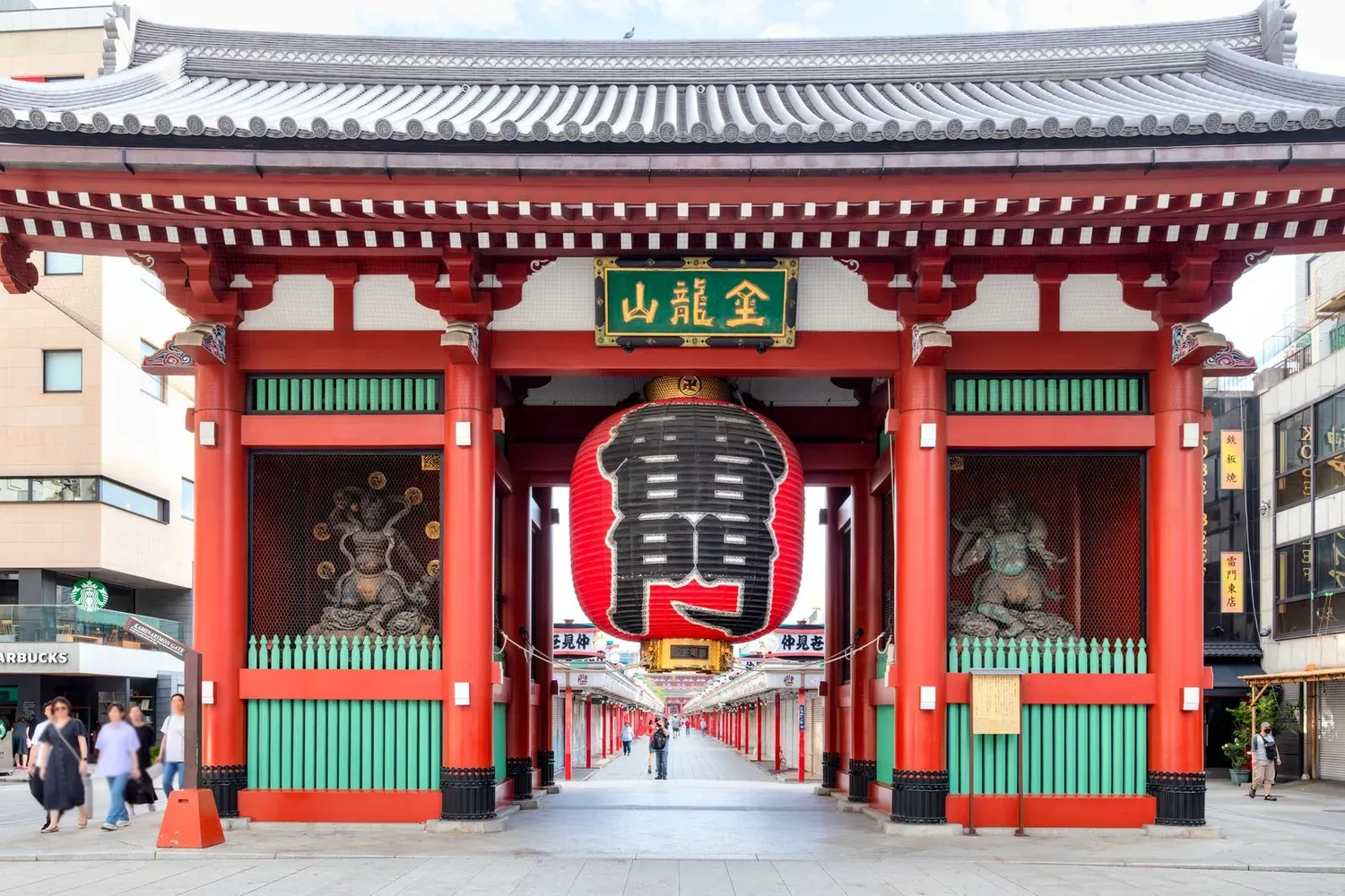 Kaminarimon gate in Asakusa with its huge red lantern and gold-lettered signboard on a bright summer morning, a few visitors walking down the Nakamise approach beyond it