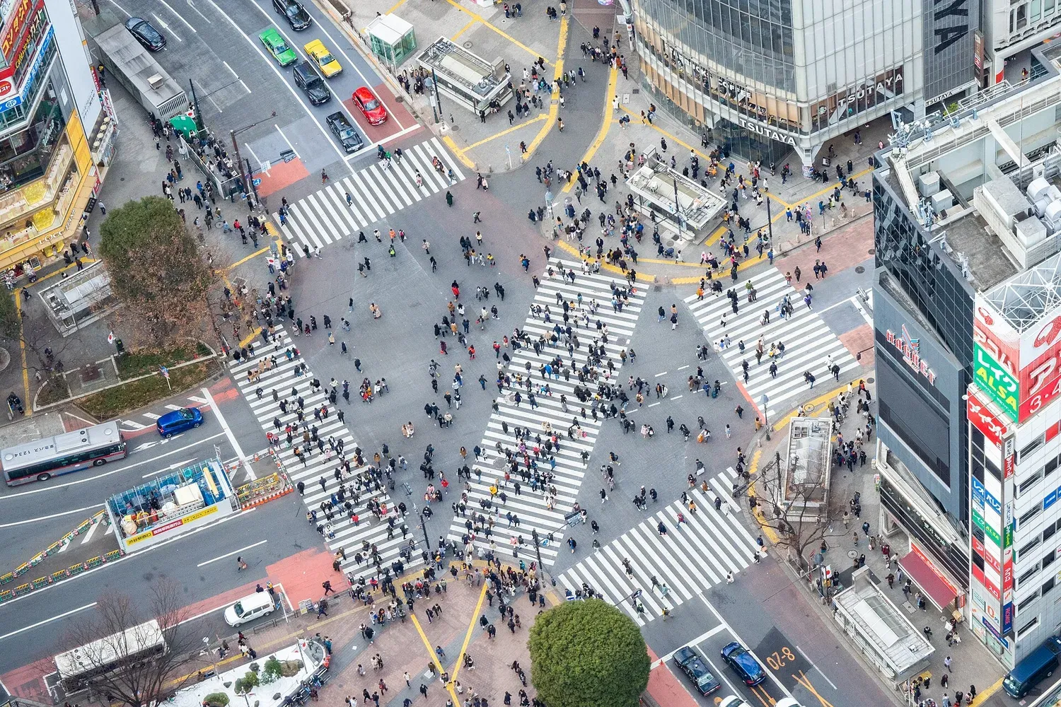 Shibuya Crossing seen from high above in daylight, with crowds crossing diagonally between tall buildings