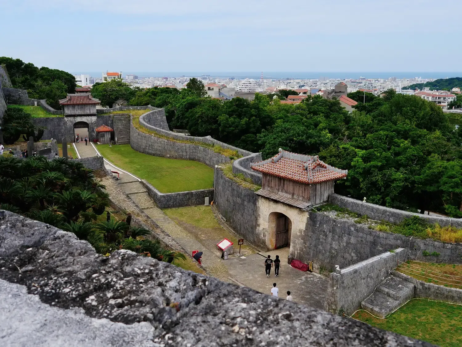 Shuri Castle seen from its walls: curving stone ramparts and gate towers above green lawns, a few visitors on the path, the city of Naha beyond