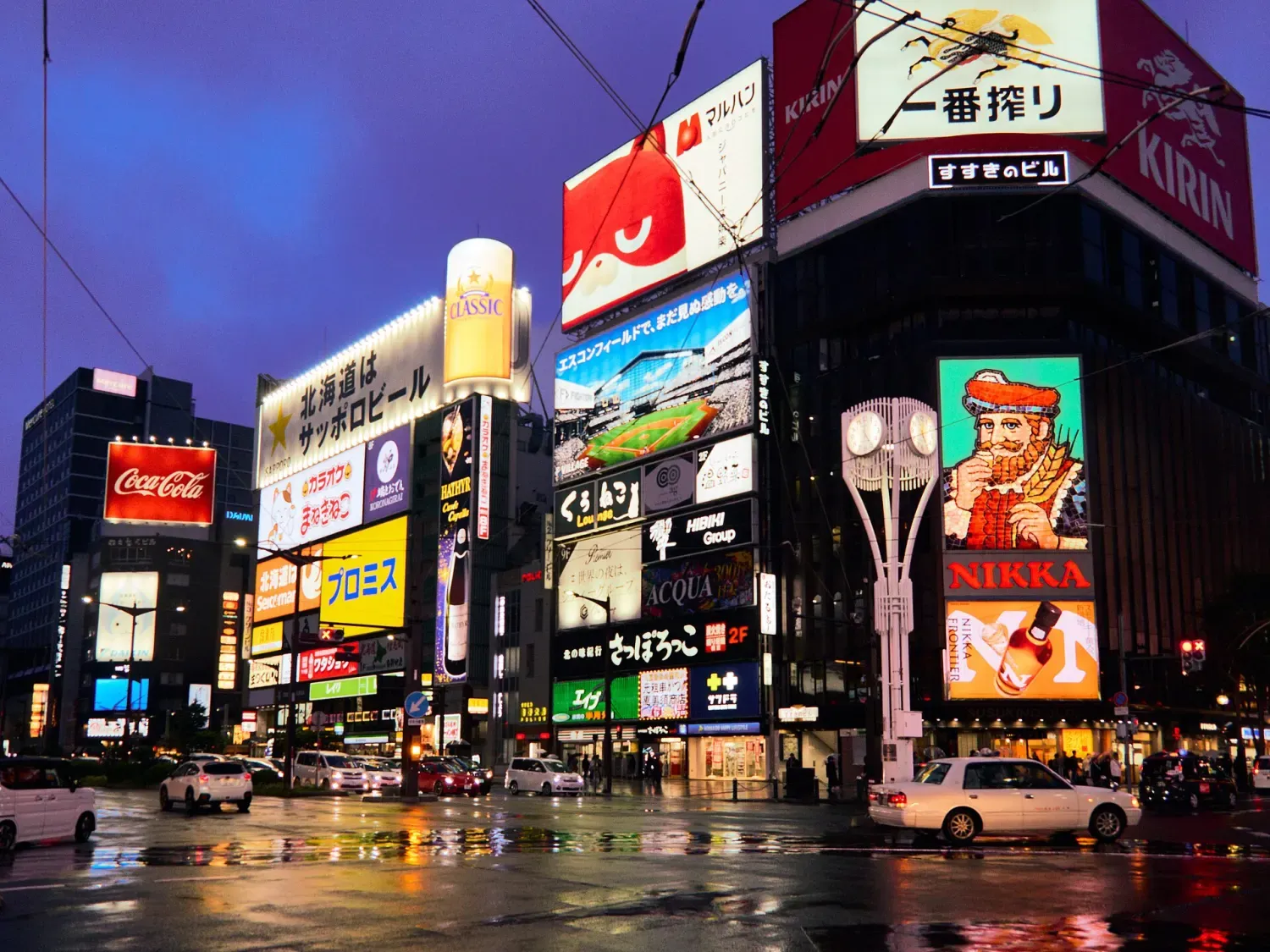 A wet intersection in Susukino at night, walls of lit signs on every building, a taxi passing below