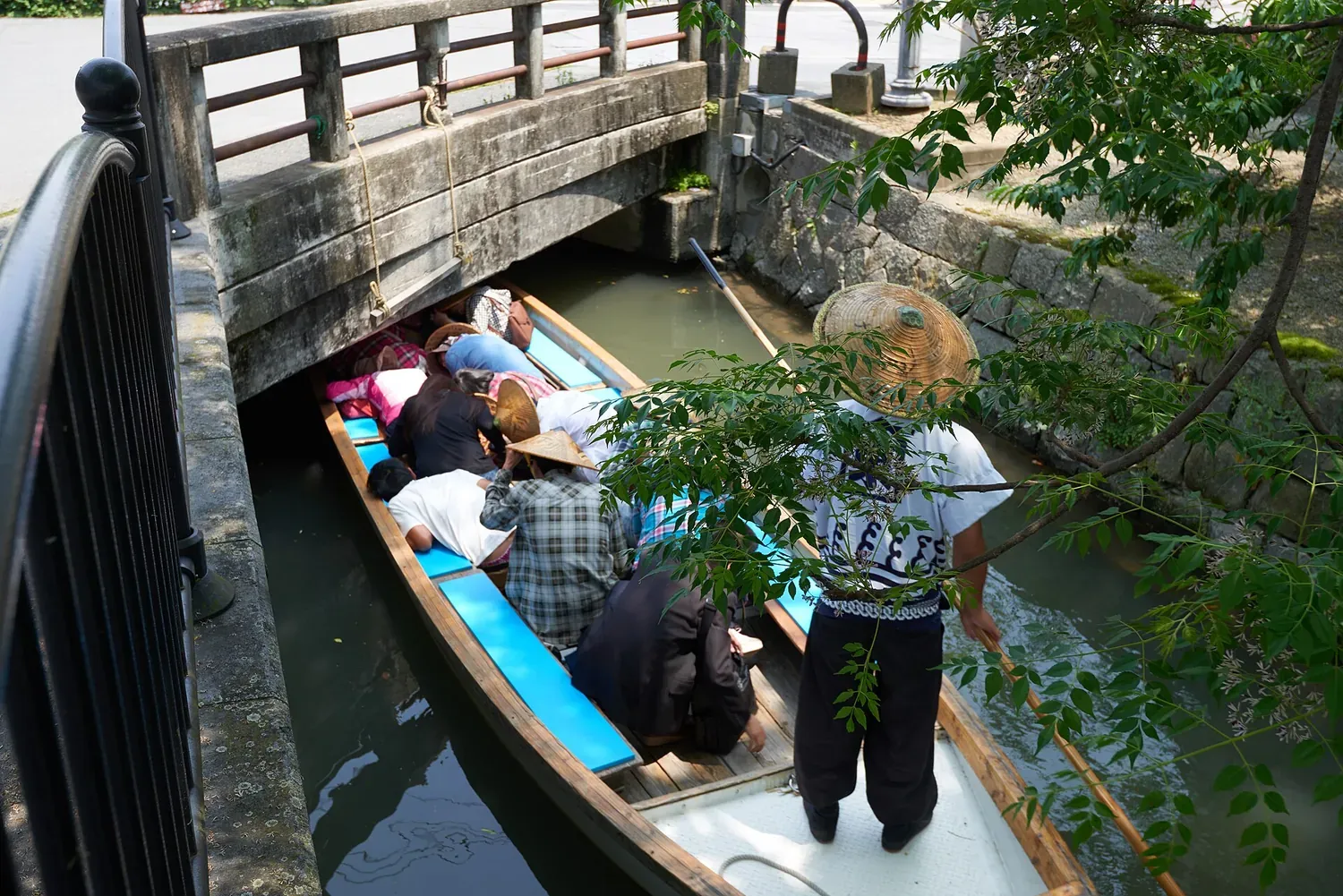 Passengers bending low in a long wooden boat as it passes under a stone bridge on a narrow canal, with the boatman standing behind them
