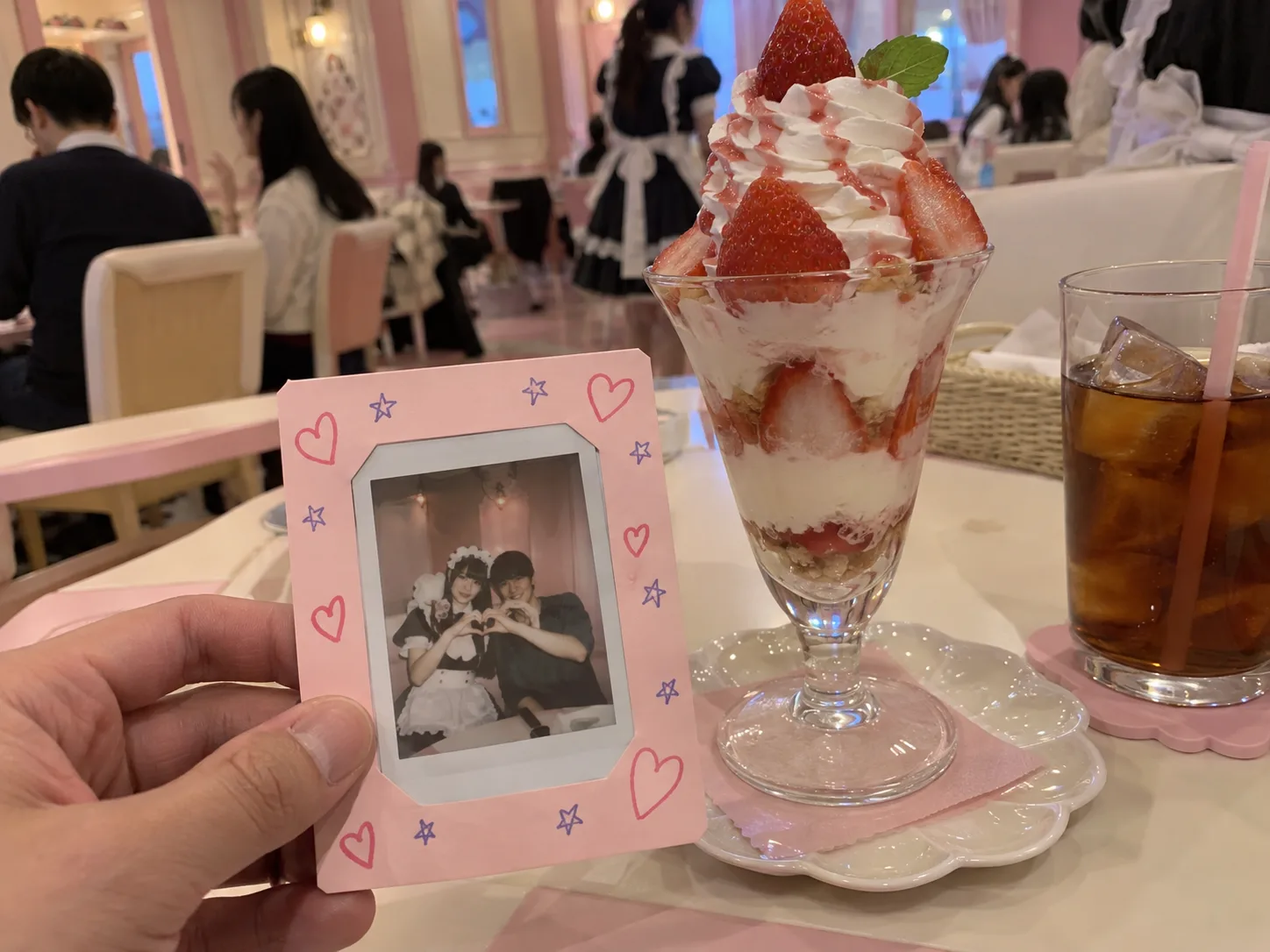An instant photo of a maid and a guest in a pastel paper mount with drawn hearts, next to a strawberry parfait