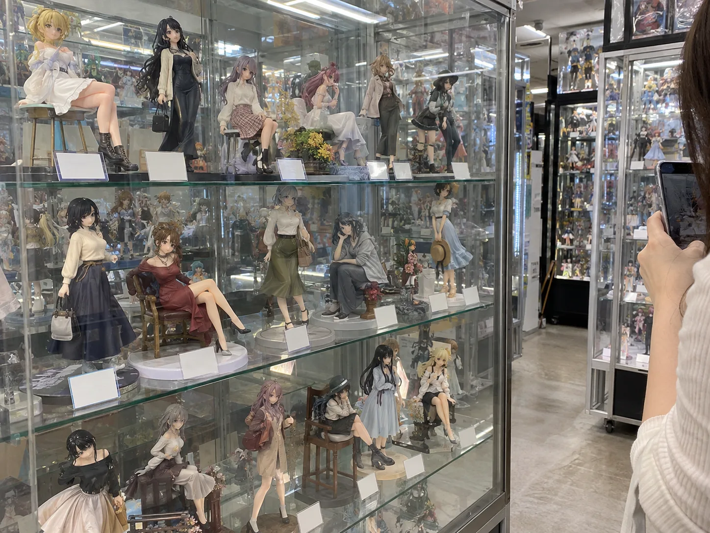 A glass display case of painted anime-style figures in a shop, with a shopper taking a phone photo at the edge