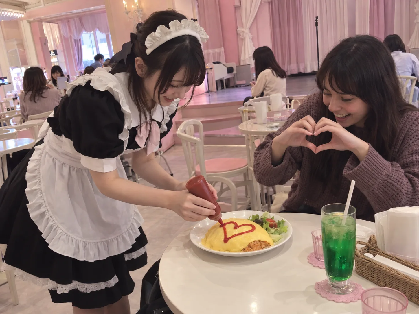 A maid piping a red heart onto an omelet rice while a guest makes a heart shape with both hands