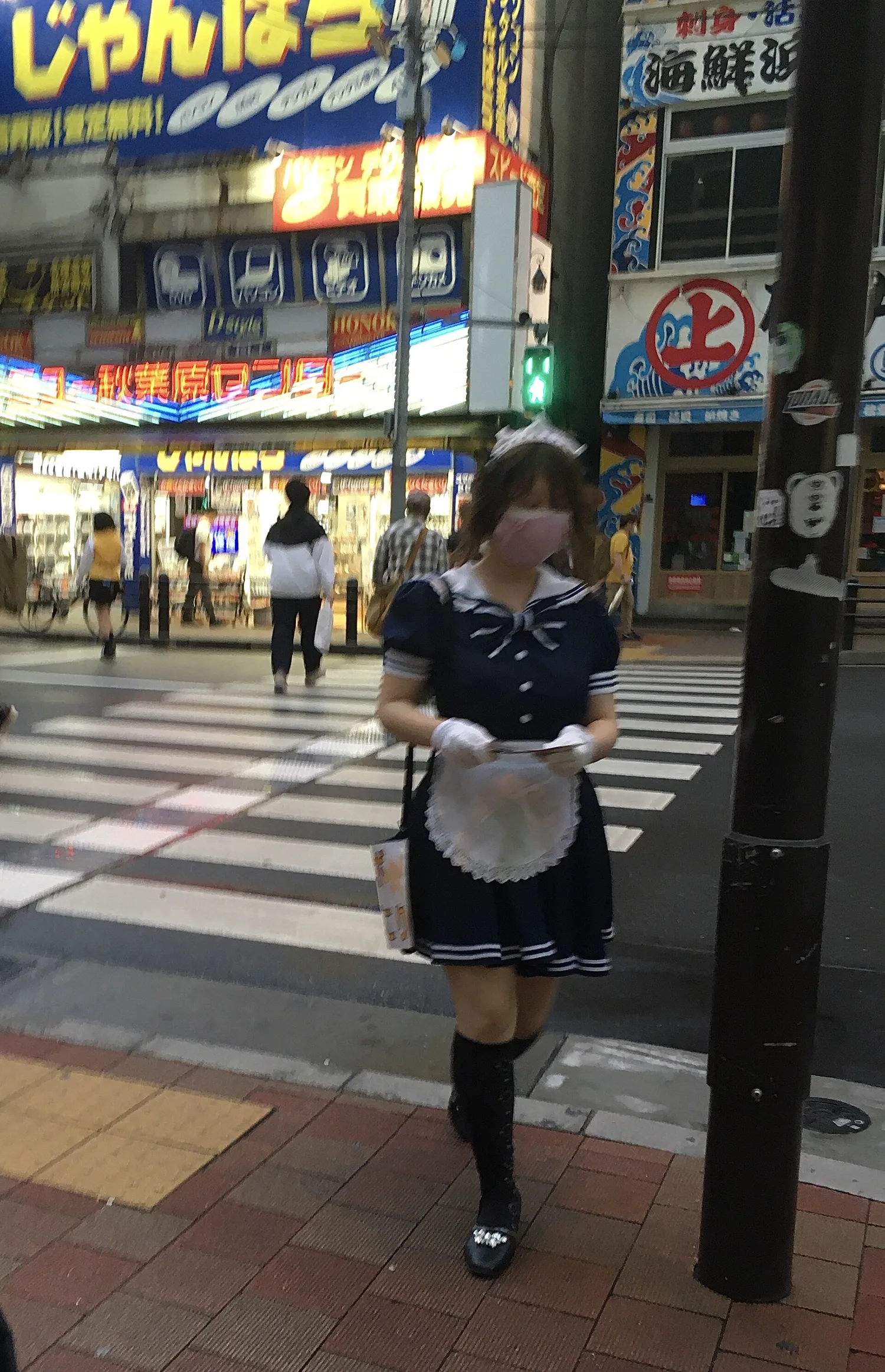 A café worker in a navy maid uniform and face mask holding flyers at an Akihabara crossing in the evening