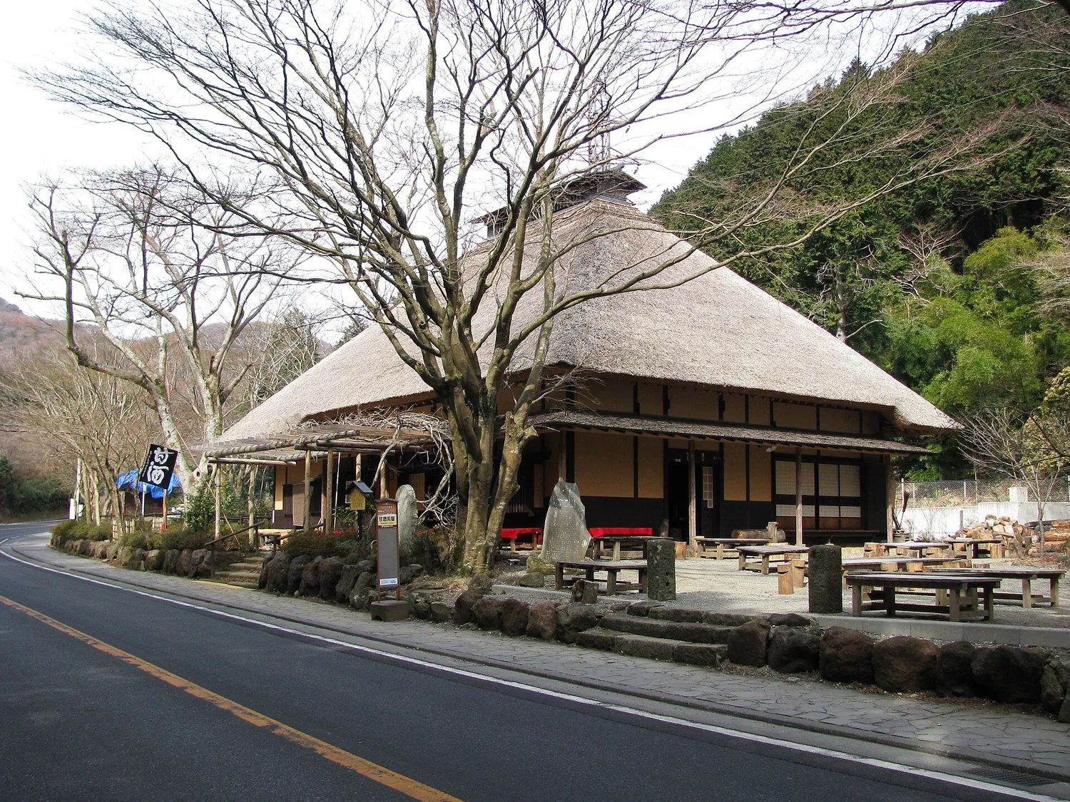 A thatched teahouse beside a road, with a bare tree in front and wooded hills behind