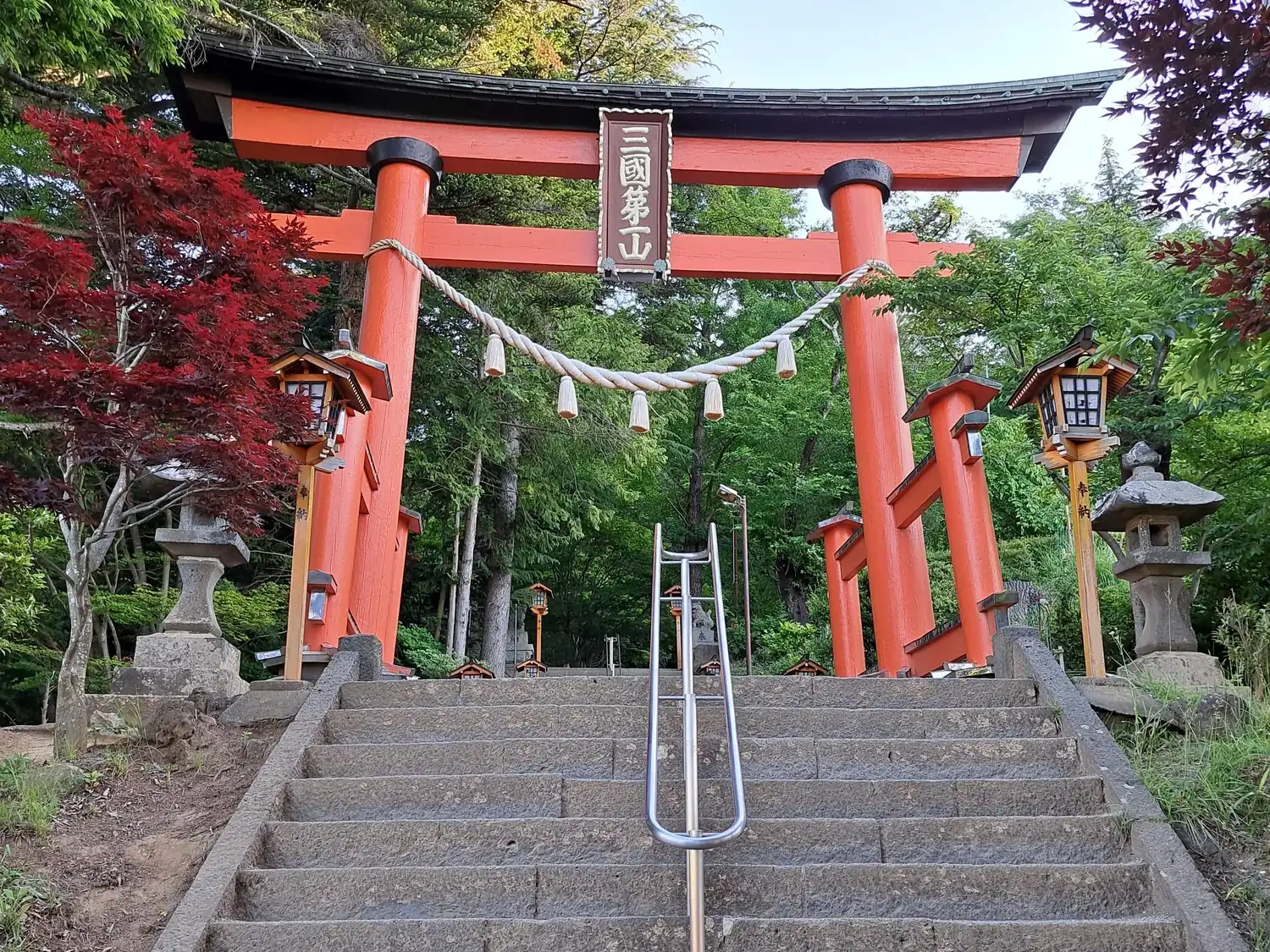 A red torii gate at the top of a flight of stone steps with a metal handrail, among green and red trees