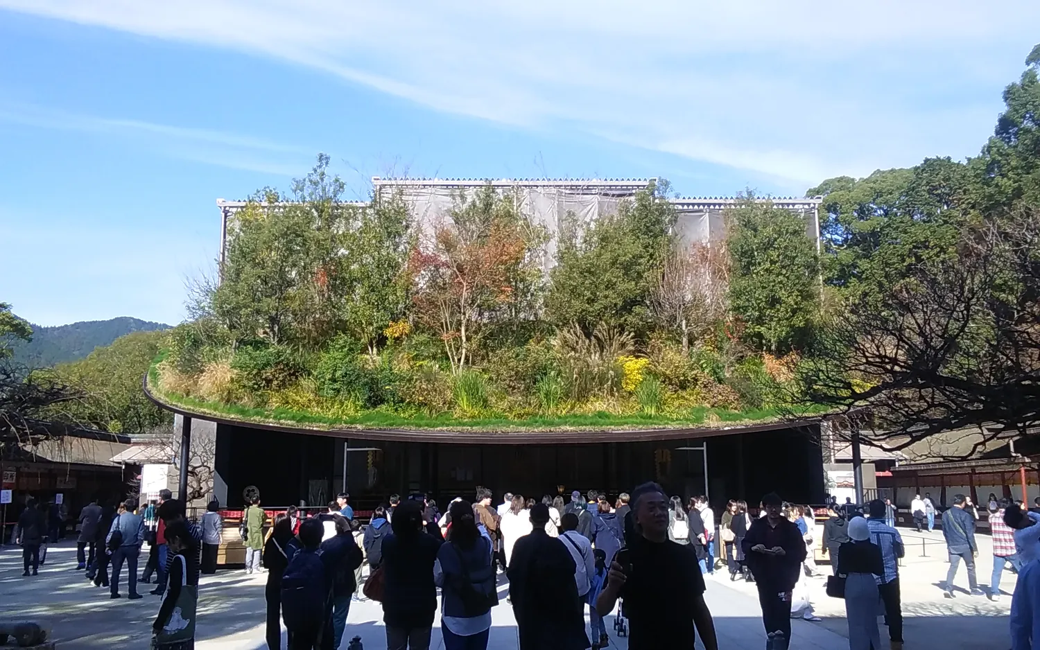 A low dark pavilion with trees and plants growing on its roof, with a crowd of visitors in front
