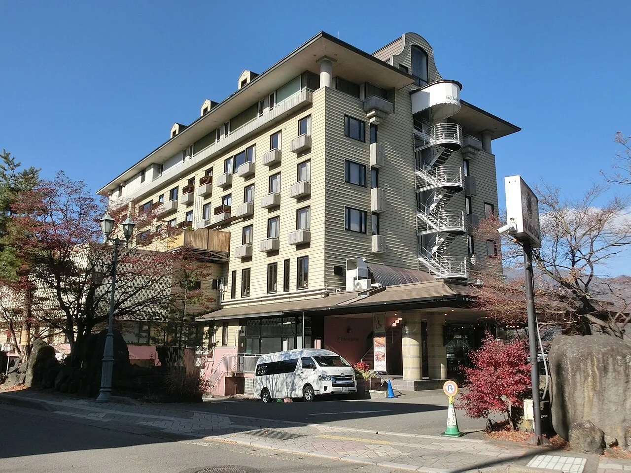 The cream-coloured, many-storeyed Fuji Lake Hotel under a clear blue sky, with a white shuttle van parked by the entrance and autumn trees in front