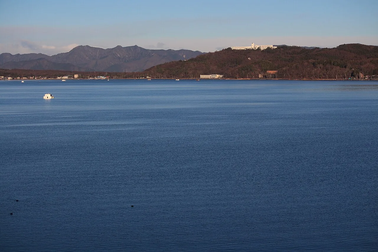 Lake Yamanaka in calm blue water, with a white hotel on the wooded hilltop across the lake and mountains behind