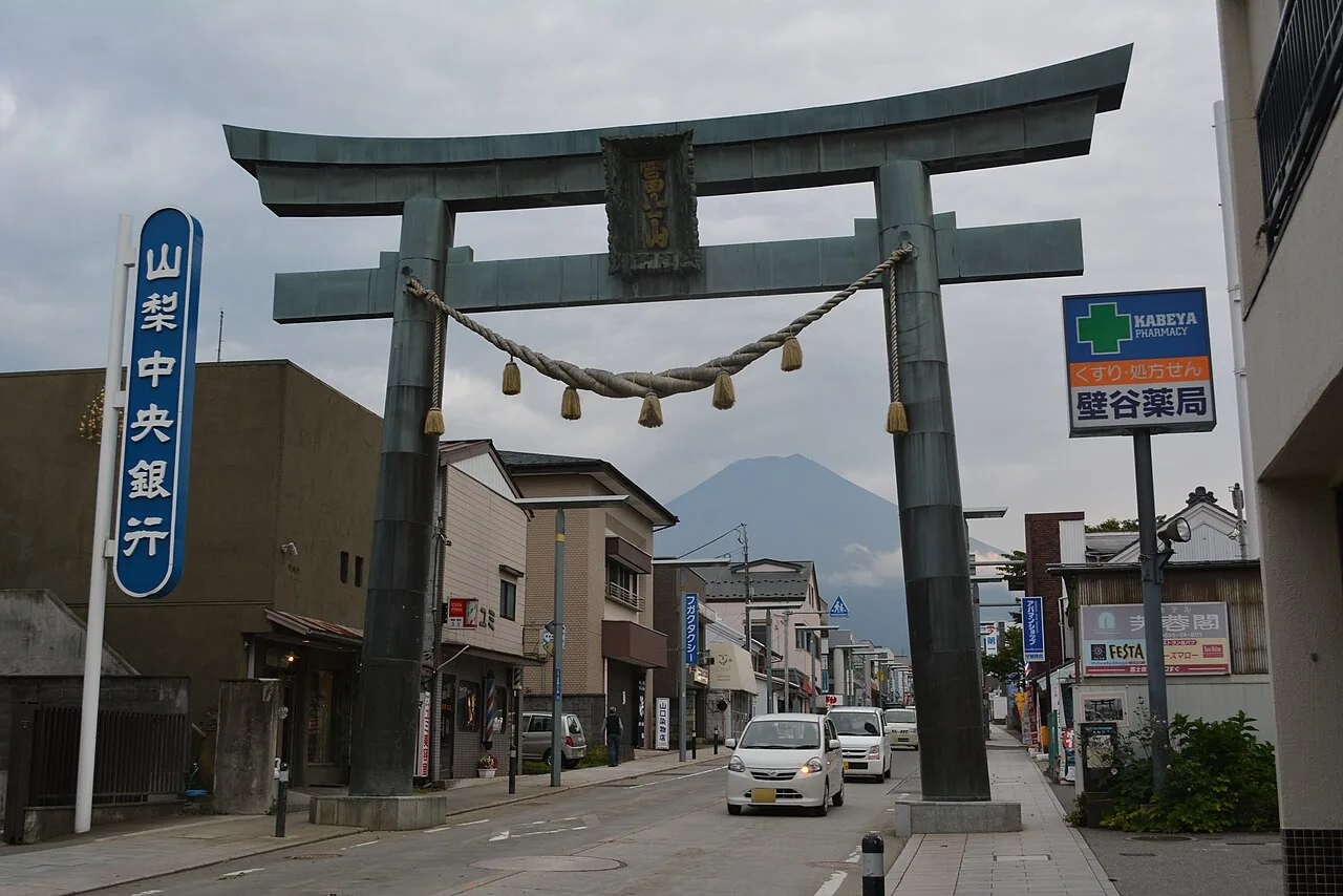 The large bronze Kanadorii gate spanning a town street in Fujiyoshida, a rope hung across it, with the grey cone of Mount Fuji in the clouds beyond