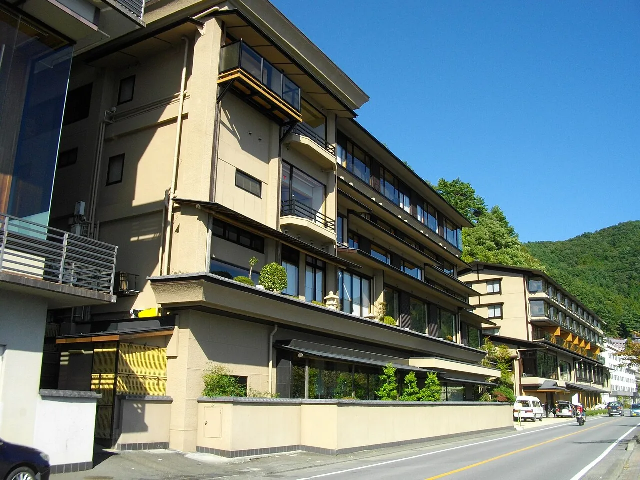 The terraced, cream-and-brown façade of Ubuya along a lakeside road, its balconies stepping back floor by floor, with green hills behind