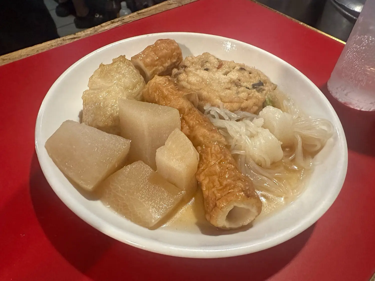 A white plate of oden on a red counter, with daikon, fish cakes and a skewer in broth
