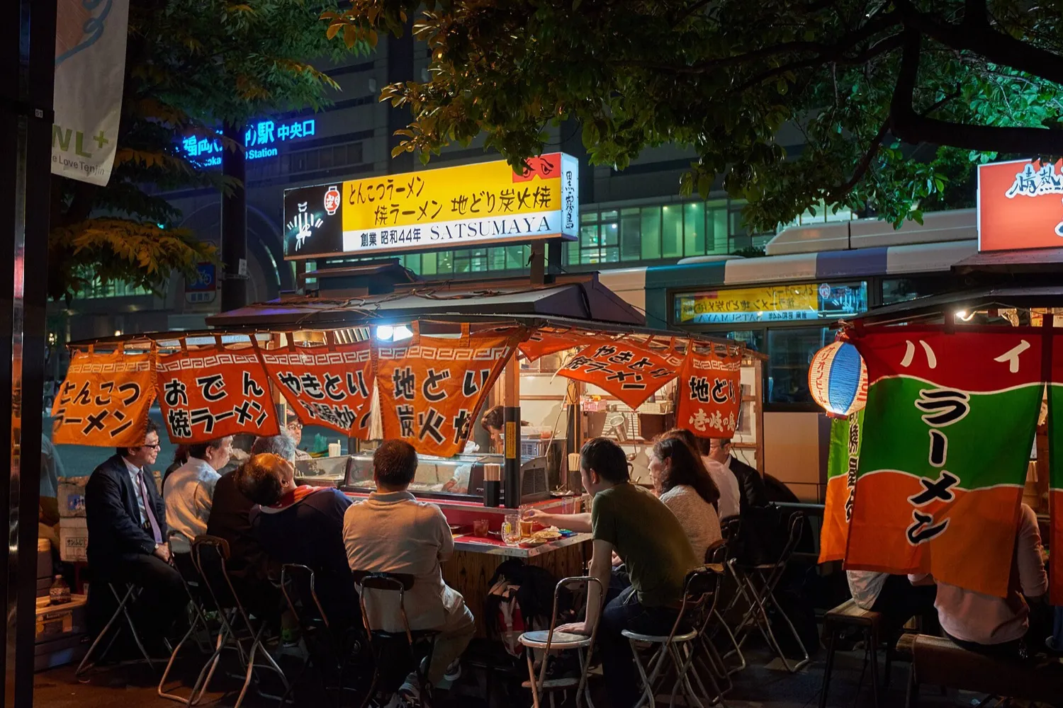 A yatai with orange curtains on a Tenjin sidewalk at night, customers on folding stools under the trees