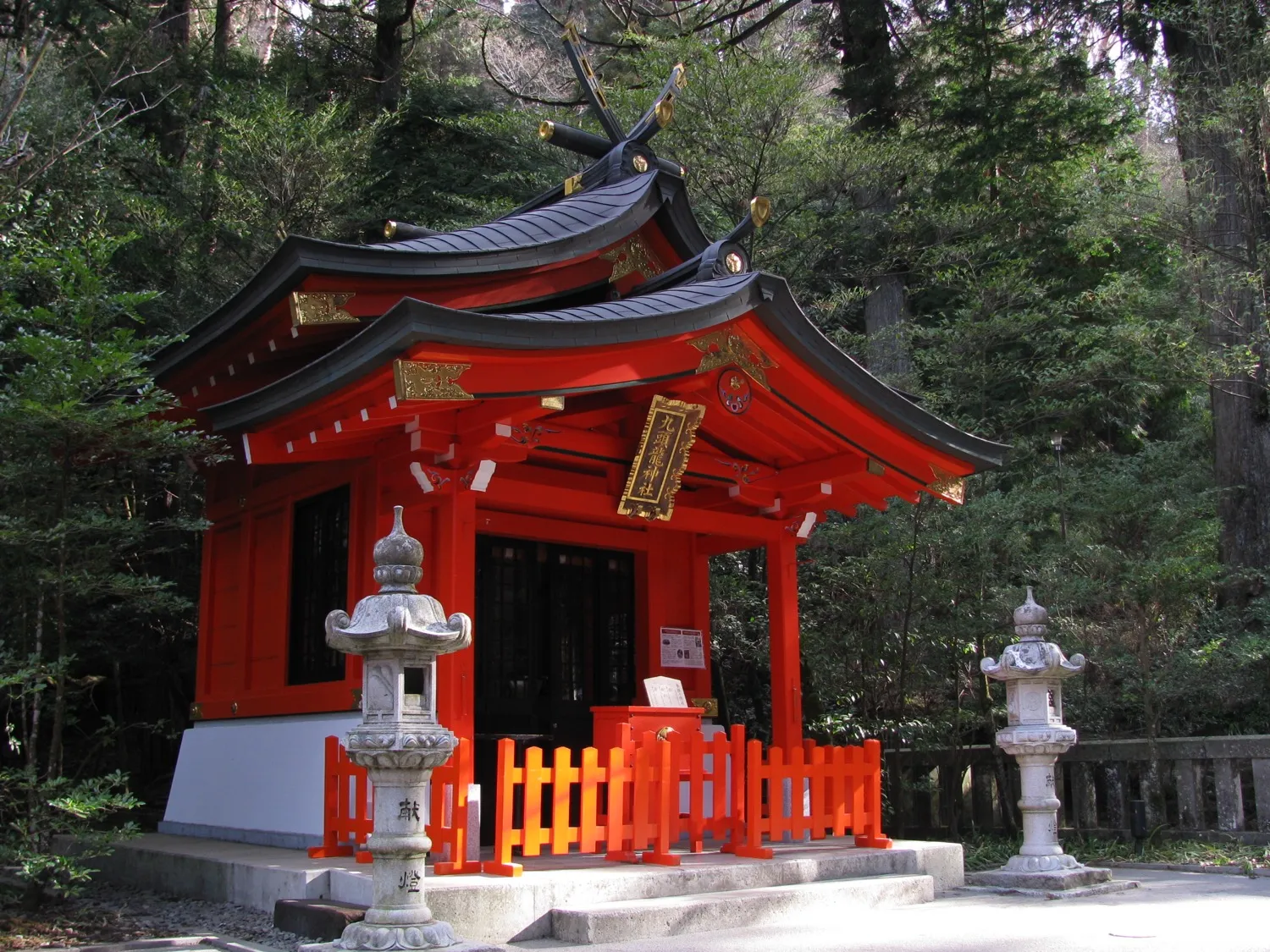 A small vermilion shrine building with stone lanterns, set among trees