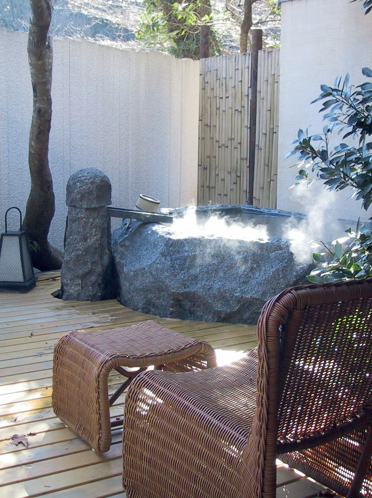 A stone tub of steaming water on a wooden deck at Gora Kadan, with wicker seats and trees behind a white wall