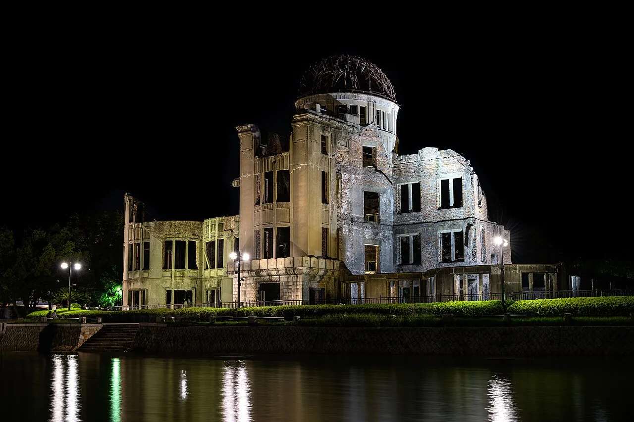 The ruined Atomic Bomb Dome lit in white at night, riverside lights reflected in the dark water below