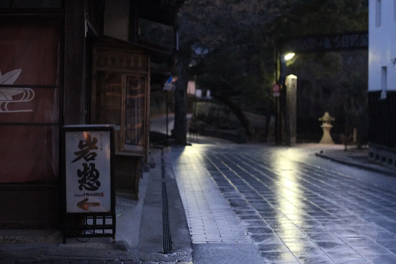 A standing sign reading Iwaso with an arrow, beside a wet stone-paved lane at dusk, a stone lantern further along the lane