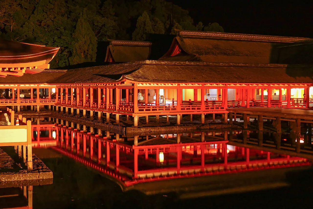 The vermilion halls and corridors of Itsukushima Shrine lit up at night, reflected in the still water beneath them