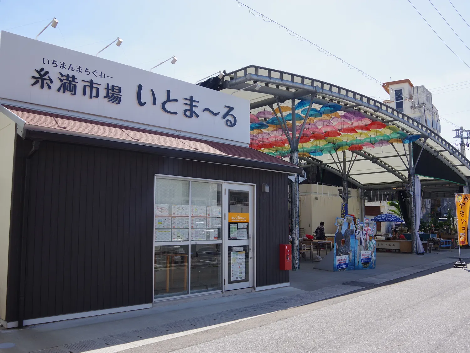 A dark wooden building with the Itomaru market name on its sign, beside a canopy hung with coloured umbrellas