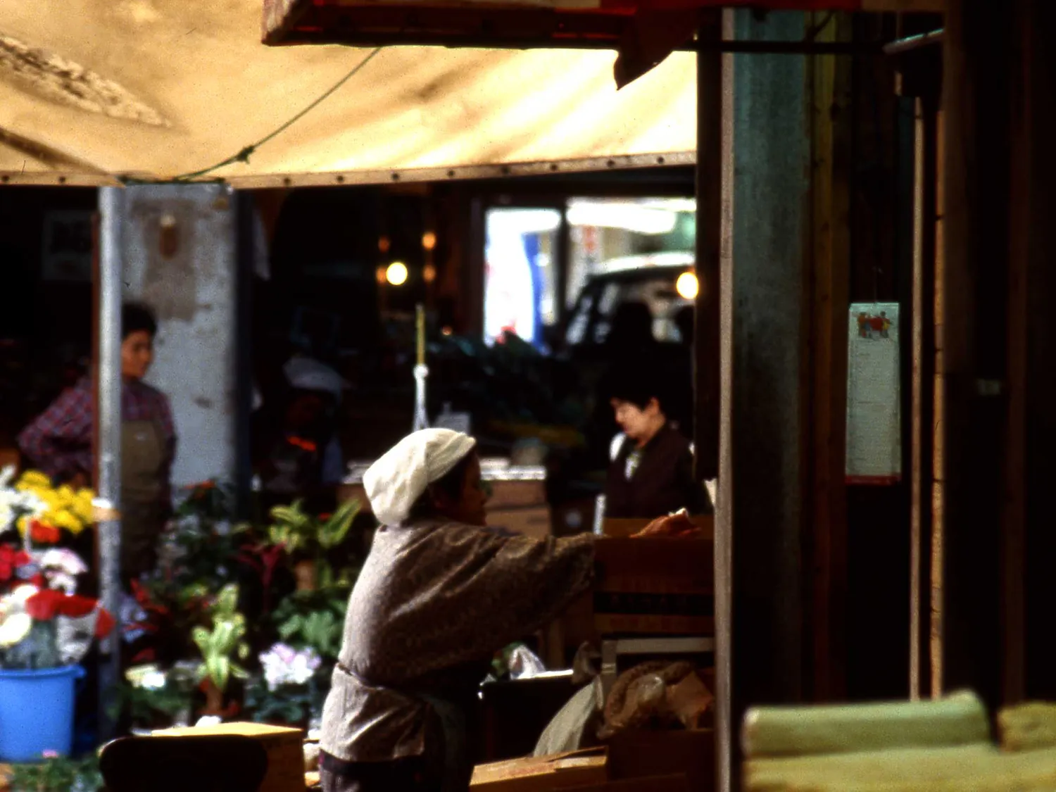 A market stall in the shade, with flowers in buckets and a seller in a white headscarf