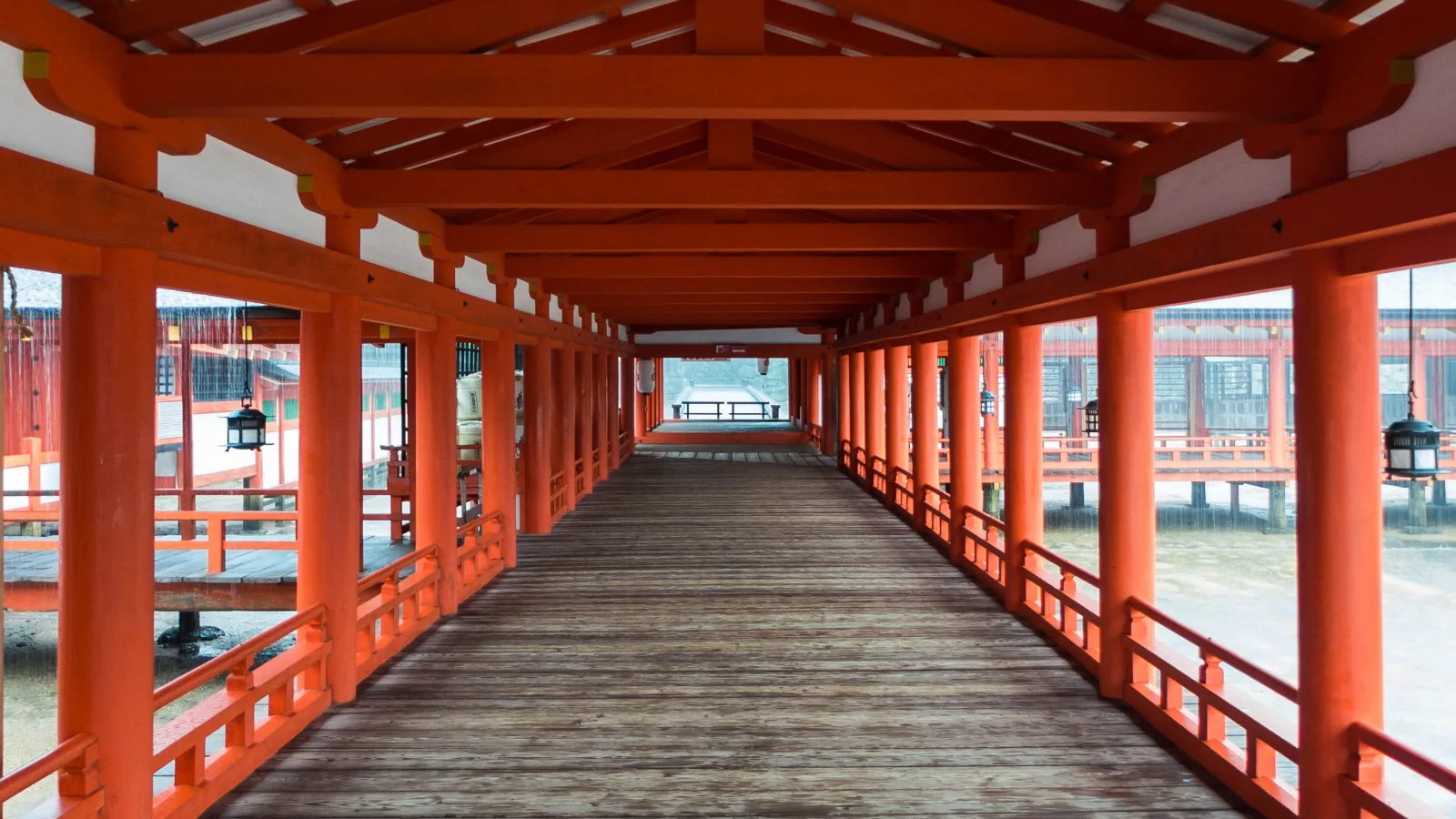 A long vermilion corridor of Itsukushima Shrine running straight ahead, lined with pillars and hanging lanterns