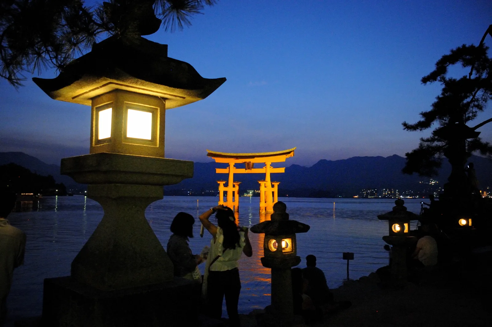 The great torii lit up at dusk, seen from the shore beside a stone lantern