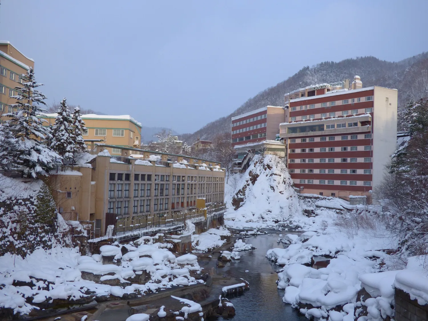Hotel buildings on both banks of a narrow river, the rocks and banks deep in snow