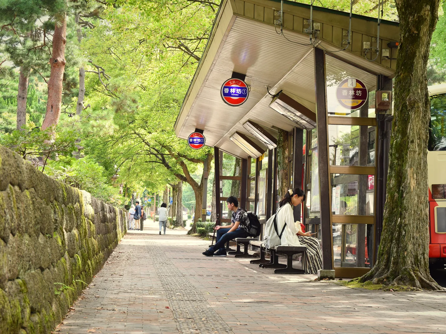 A tree-lined pavement beside an old mossy stone wall in central Kanazawa, a bus shelter and people waiting