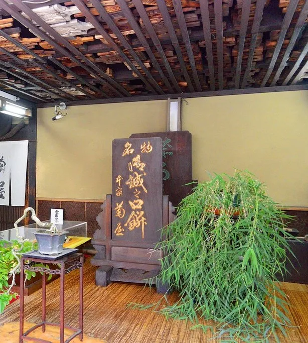 A shop interior with wooden sweet molds lining the ceiling, a tall signboard with gold lettering, and a potted bamboo