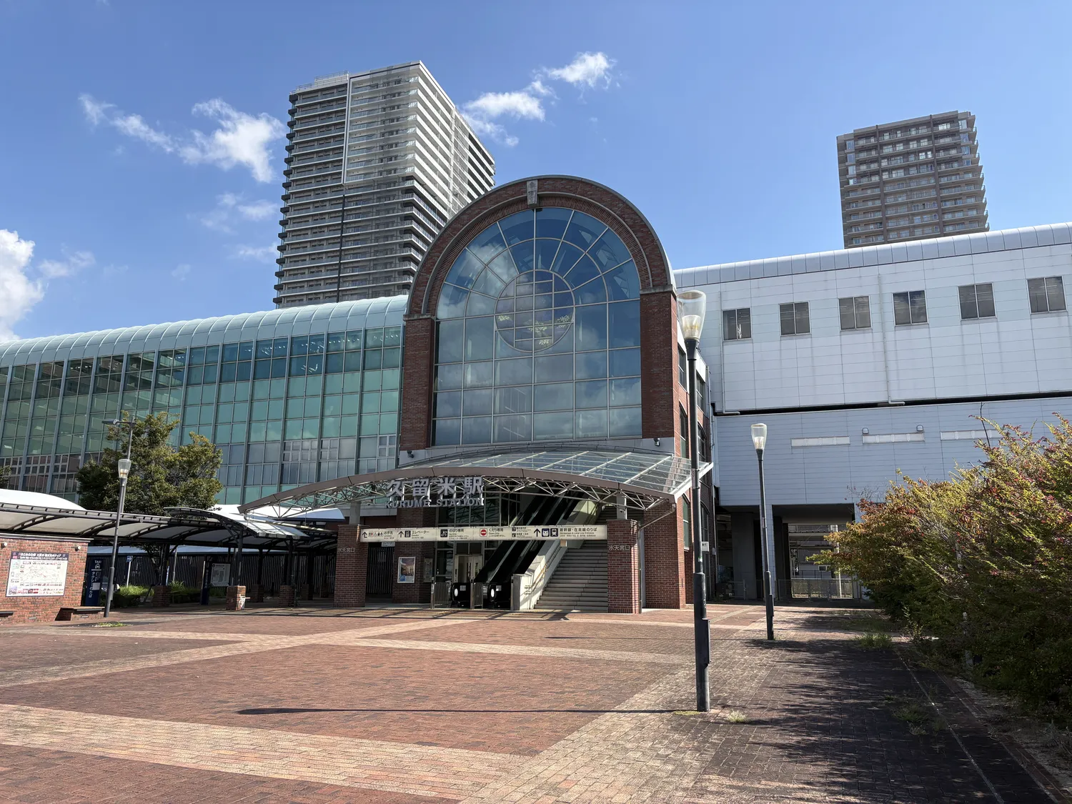 The glass-fronted entrance of a modern station building under a blue sky, with tall towers behind