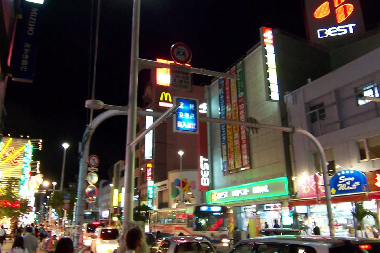 Kokusai Street at night, shop signs and neon lit along both sides, cars and people below