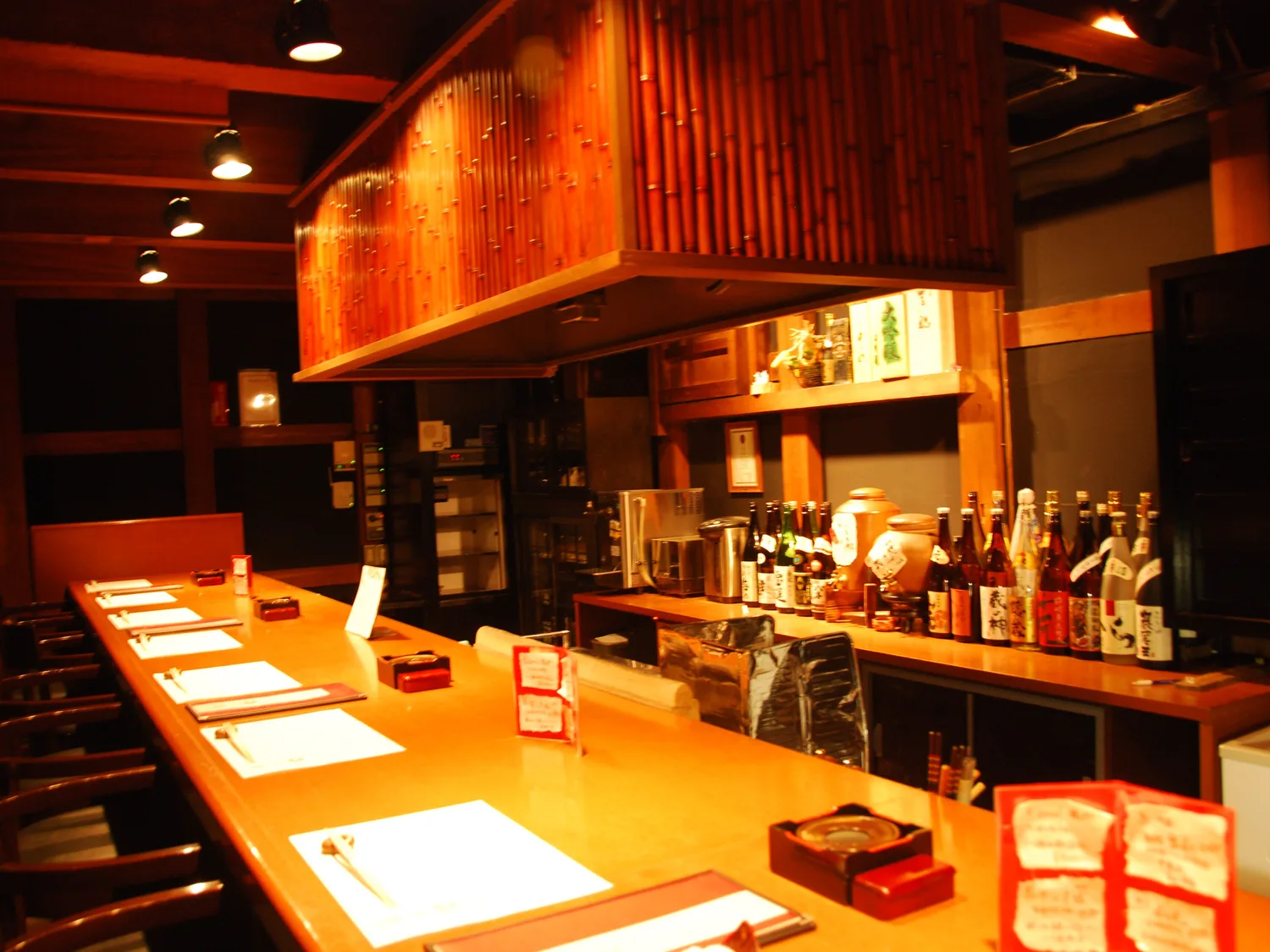 A warmly lit wooden counter with menus laid out, shelves of sake bottles behind it