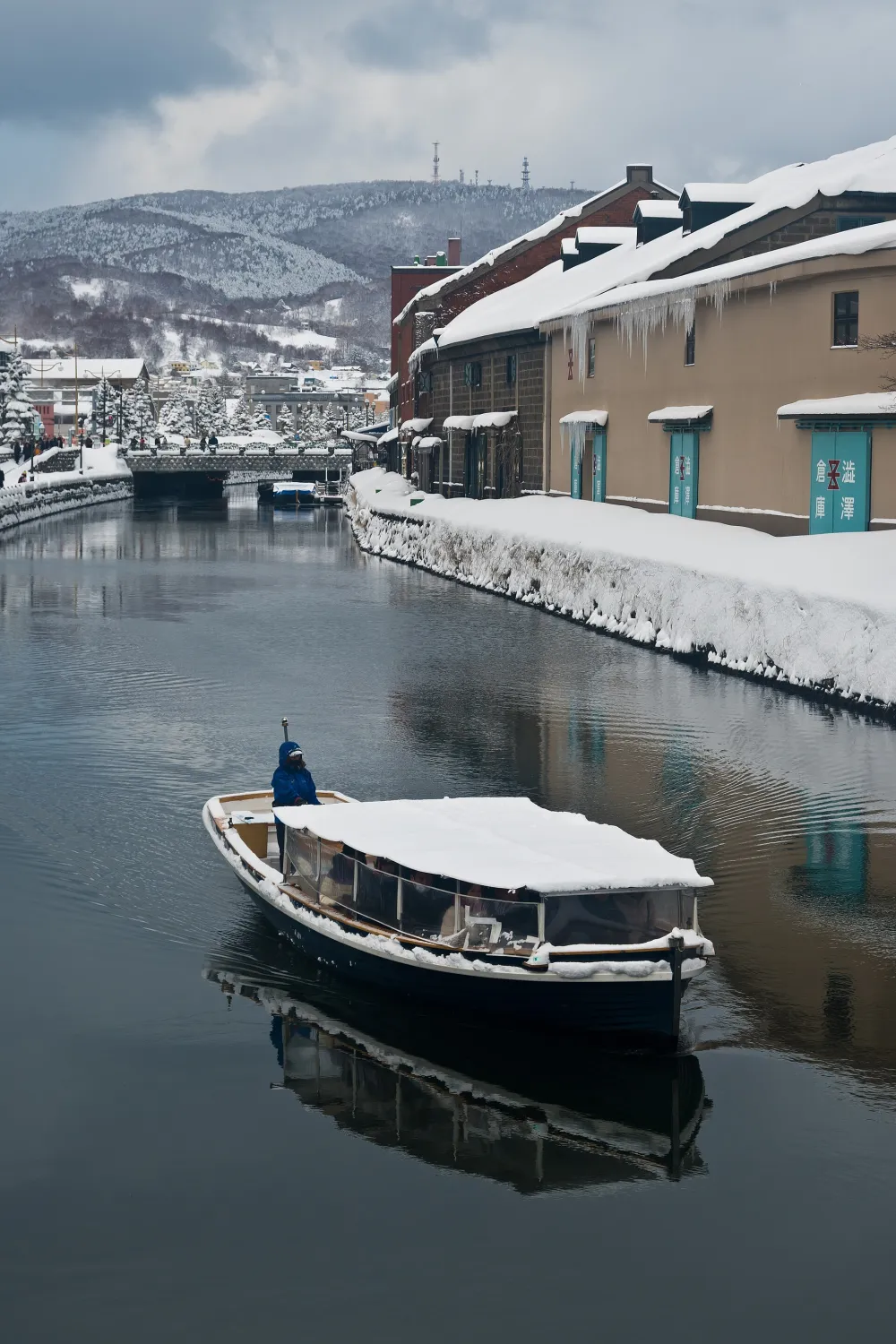 A small covered tour boat on a still canal lined with snow-covered warehouses