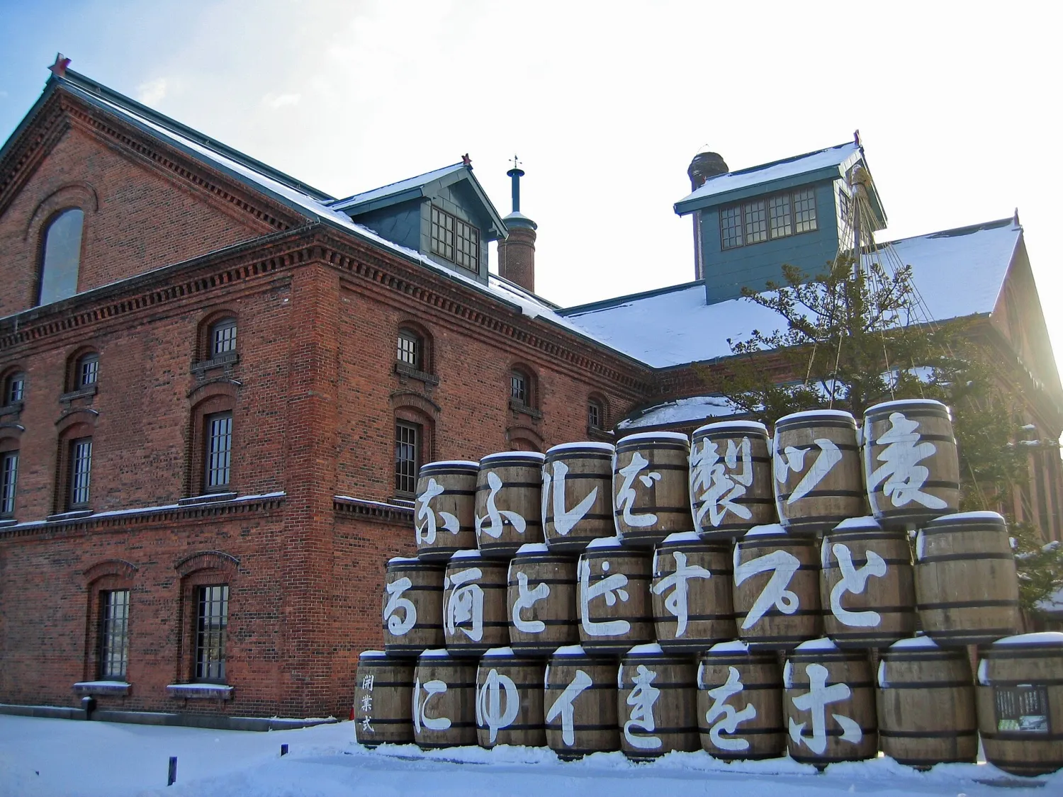 Stacked wooden barrels painted with Japanese characters in front of a snowy red-brick building